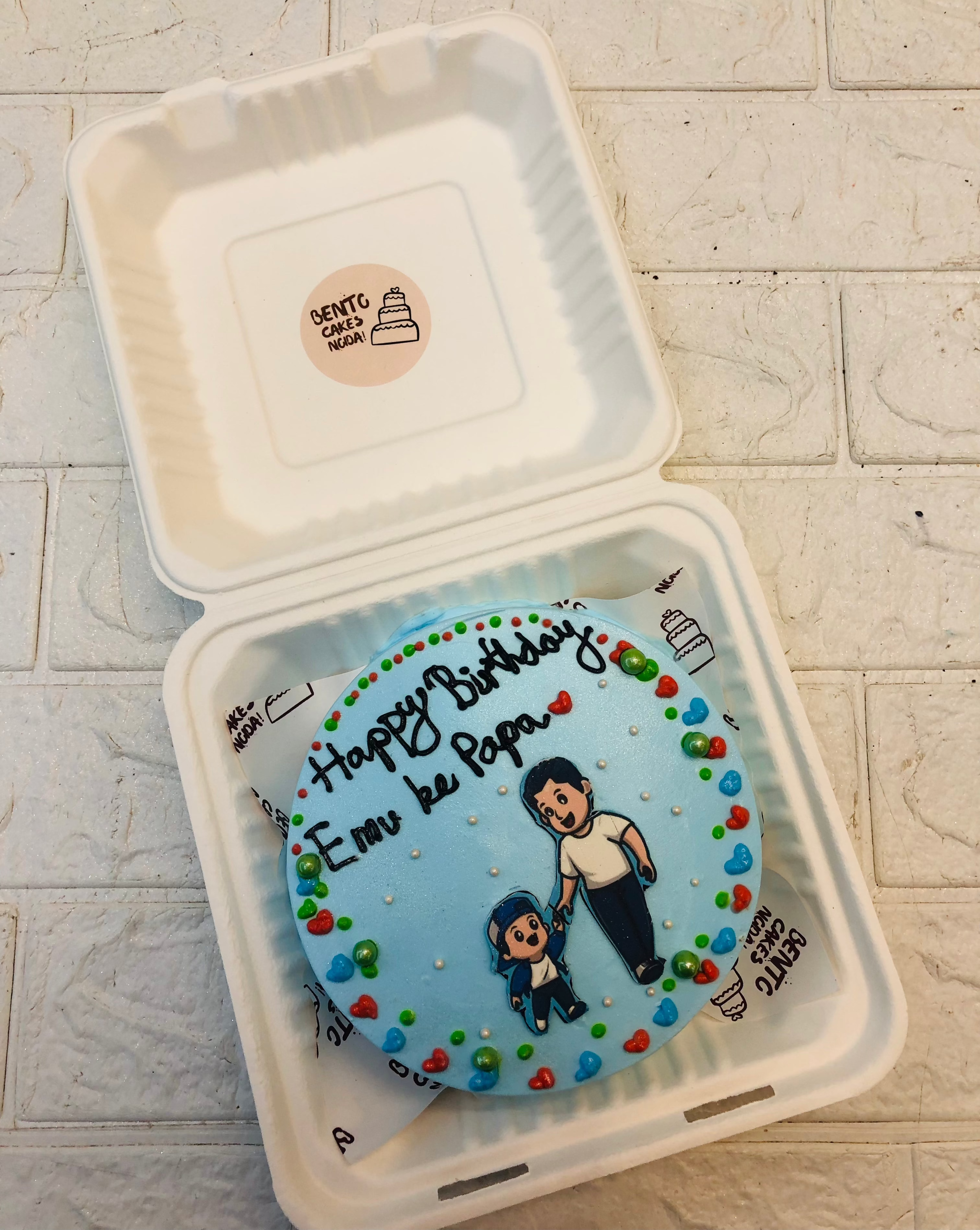 A beautifully crafted round blue birthday cake placed inside a white box, featuring an illustration of a father holding hands with a child. The cake is decorated with colorful dots and has the heartfelt message "Happy Birthday Ema ke Papa" written on it.