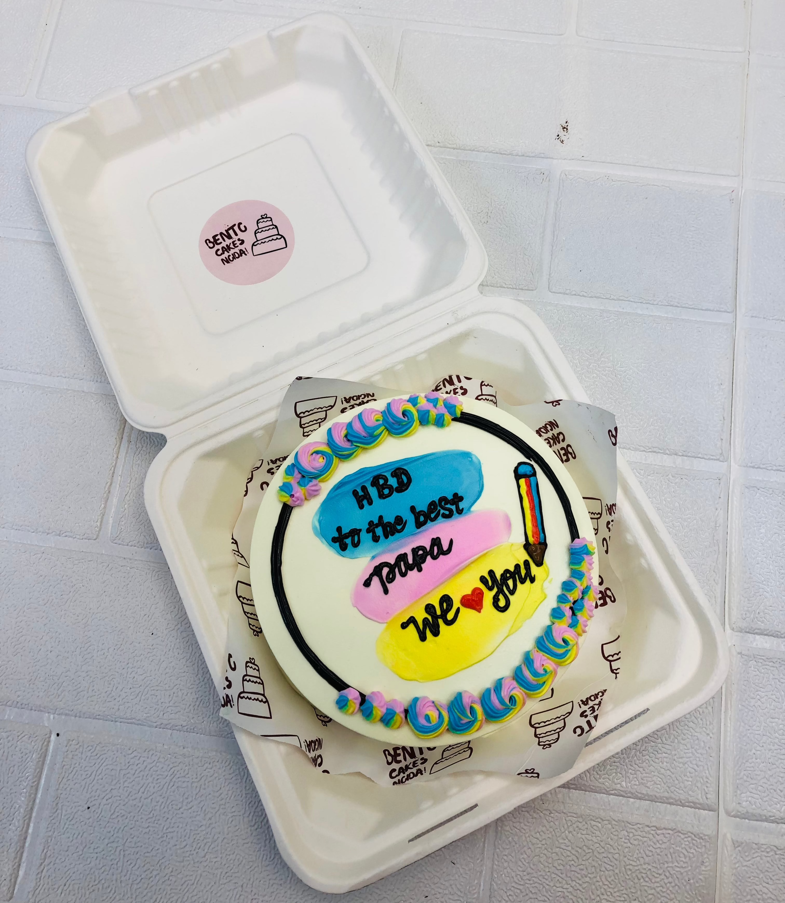 A round bento cake inside a white box, decorated with pastel swirls and a colorful birthday message saying, "HBD to the best papa, we love you."