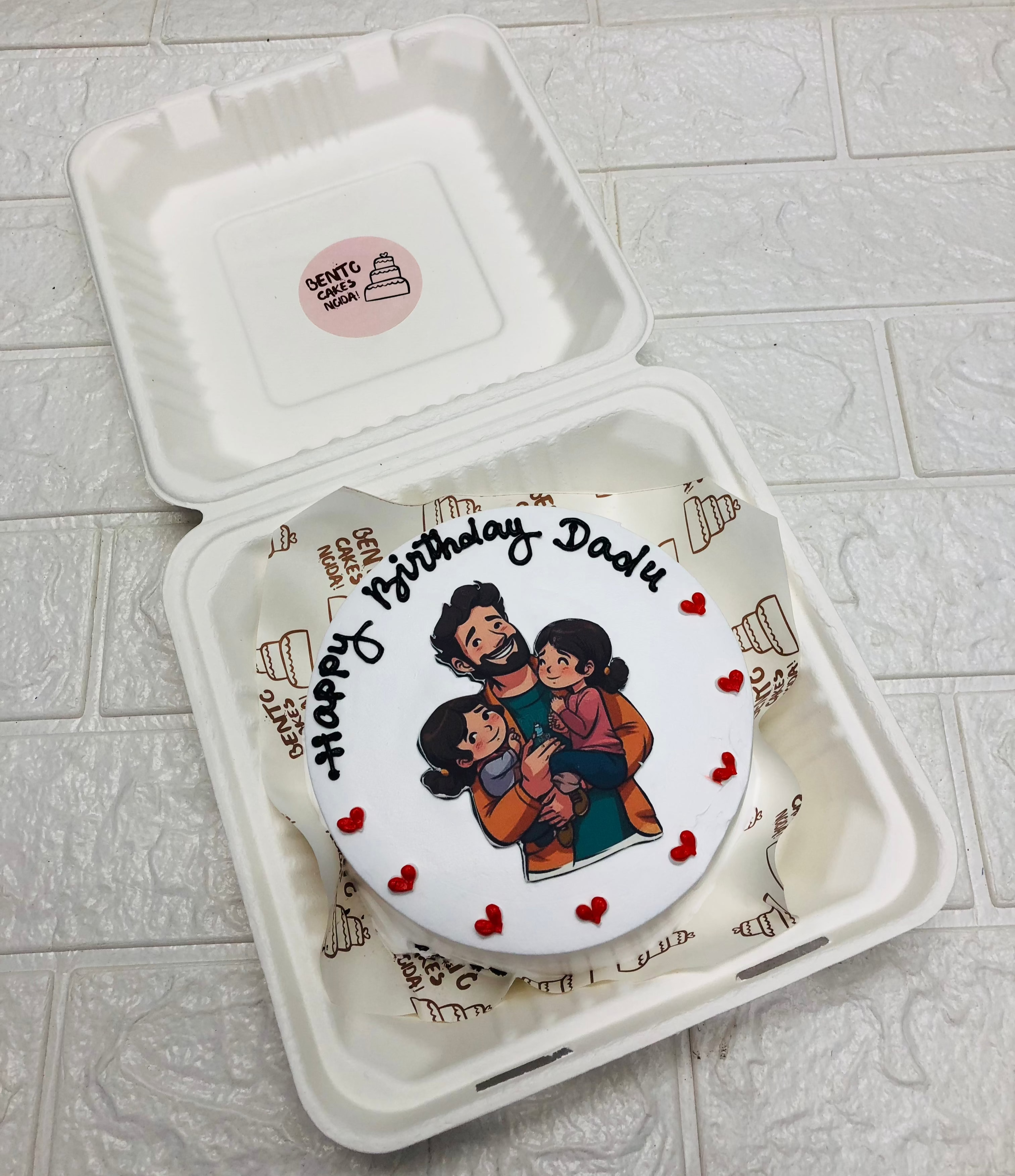 A white bento cake is designed with a father with his 2 daughters printed on cake and decorated with red hearts.