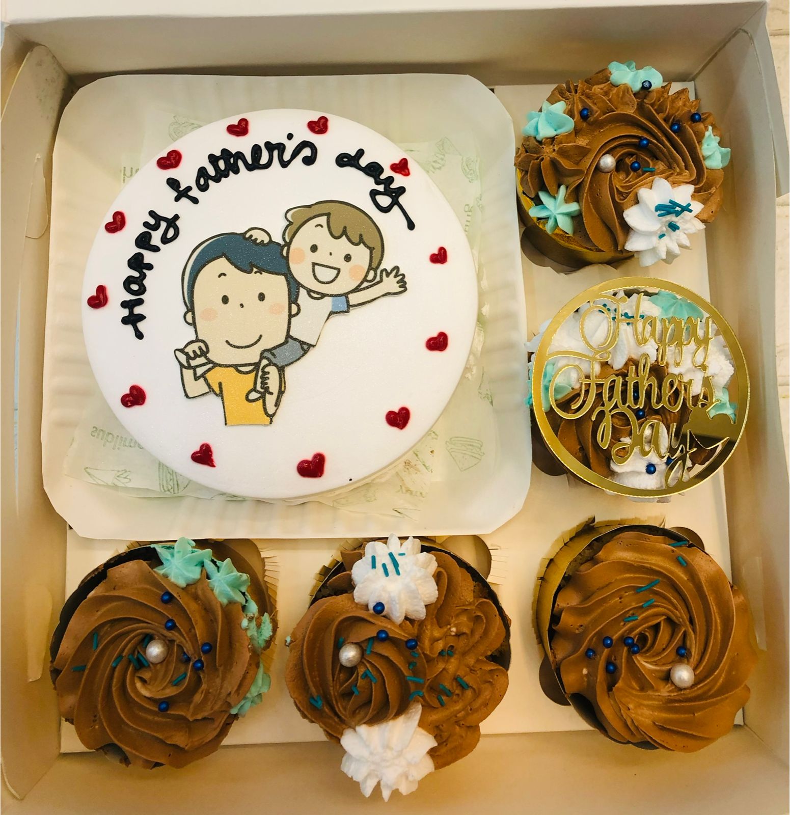 A white bento cake with fathers day carton print and 5 cupcakes with brown and white floral design.