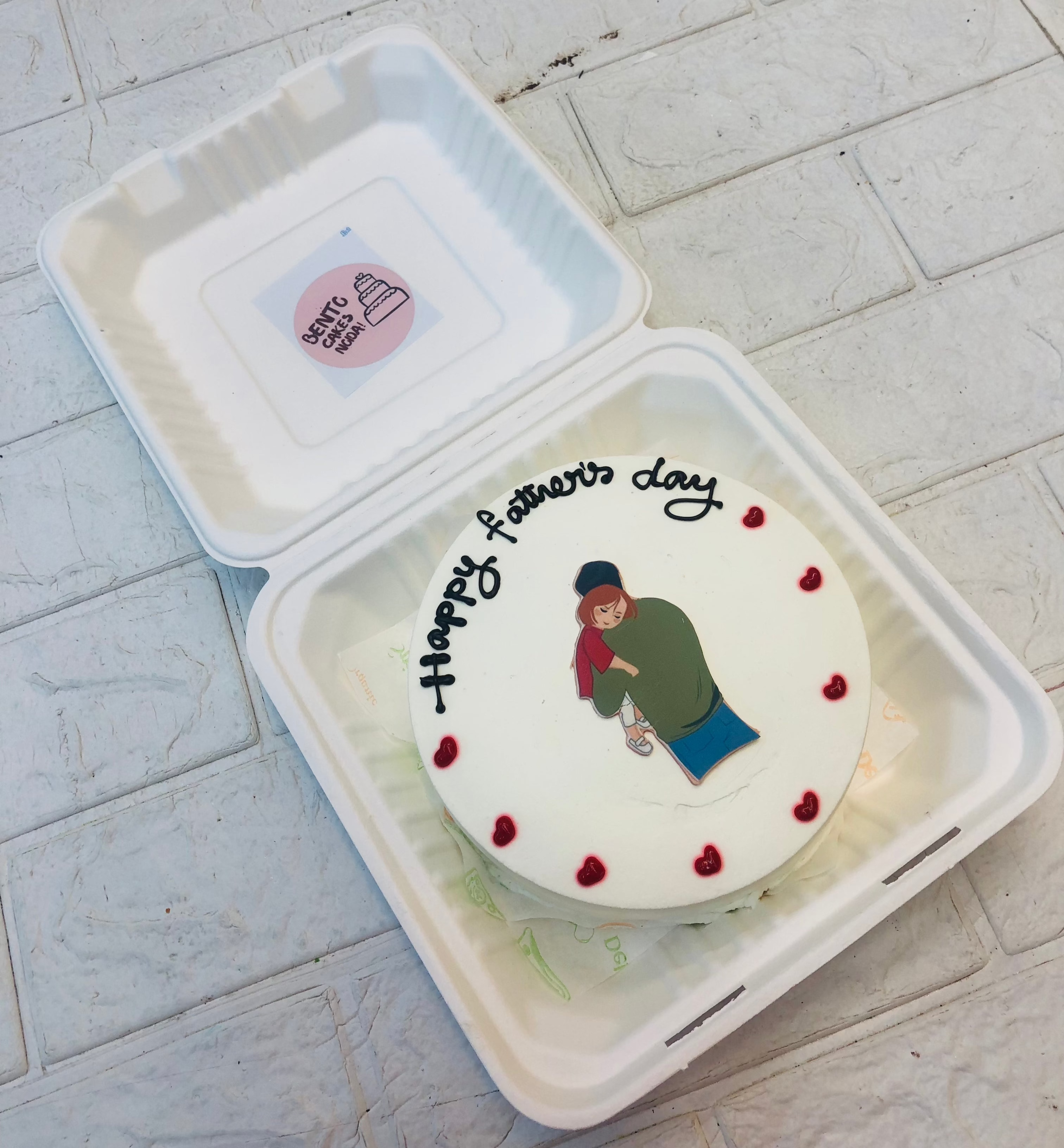 A bento cake with white icing and a print of father and kid placed on it and decorated with red hearts.