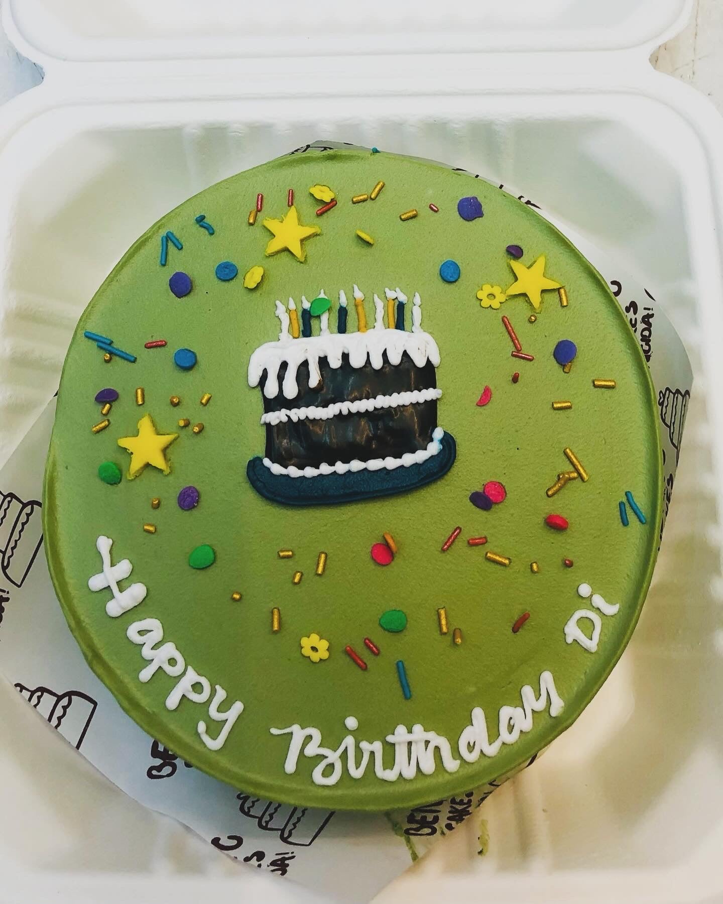 A beautifully decorated green birthday cake featuring stars, sprinkles, and a small cake illustration with candles. The cake has "Happy Birthday Di" written in white icing, adding a personal touch to the celebration.