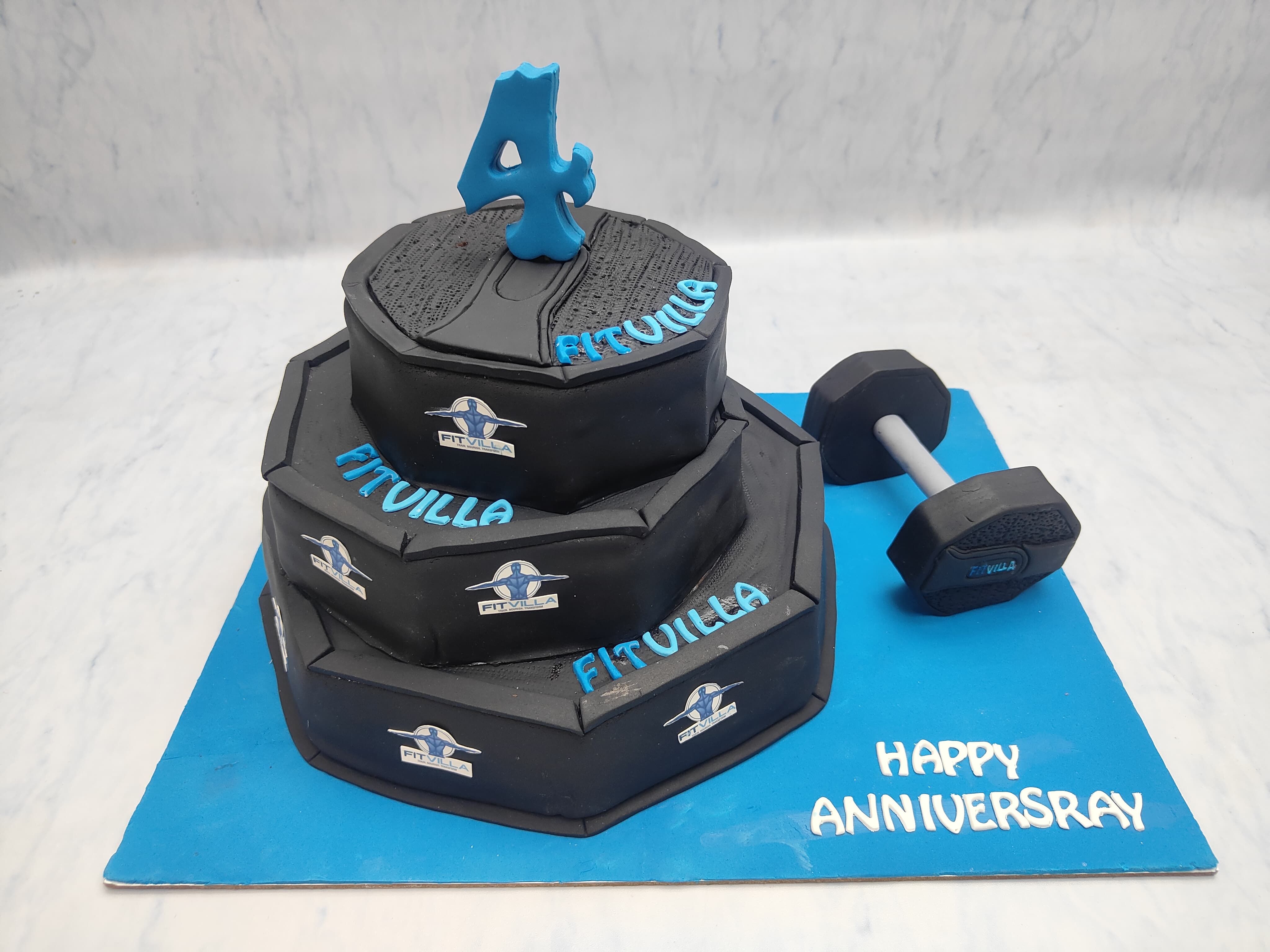A three-tiered black cake with blue accents, designed to resemble weight plates, featuring the Fitvilla logo and a blue number 4 topper. The cake is placed on a blue base with the text 'Happy Anniversary' and is accompanied by a dumbbell.