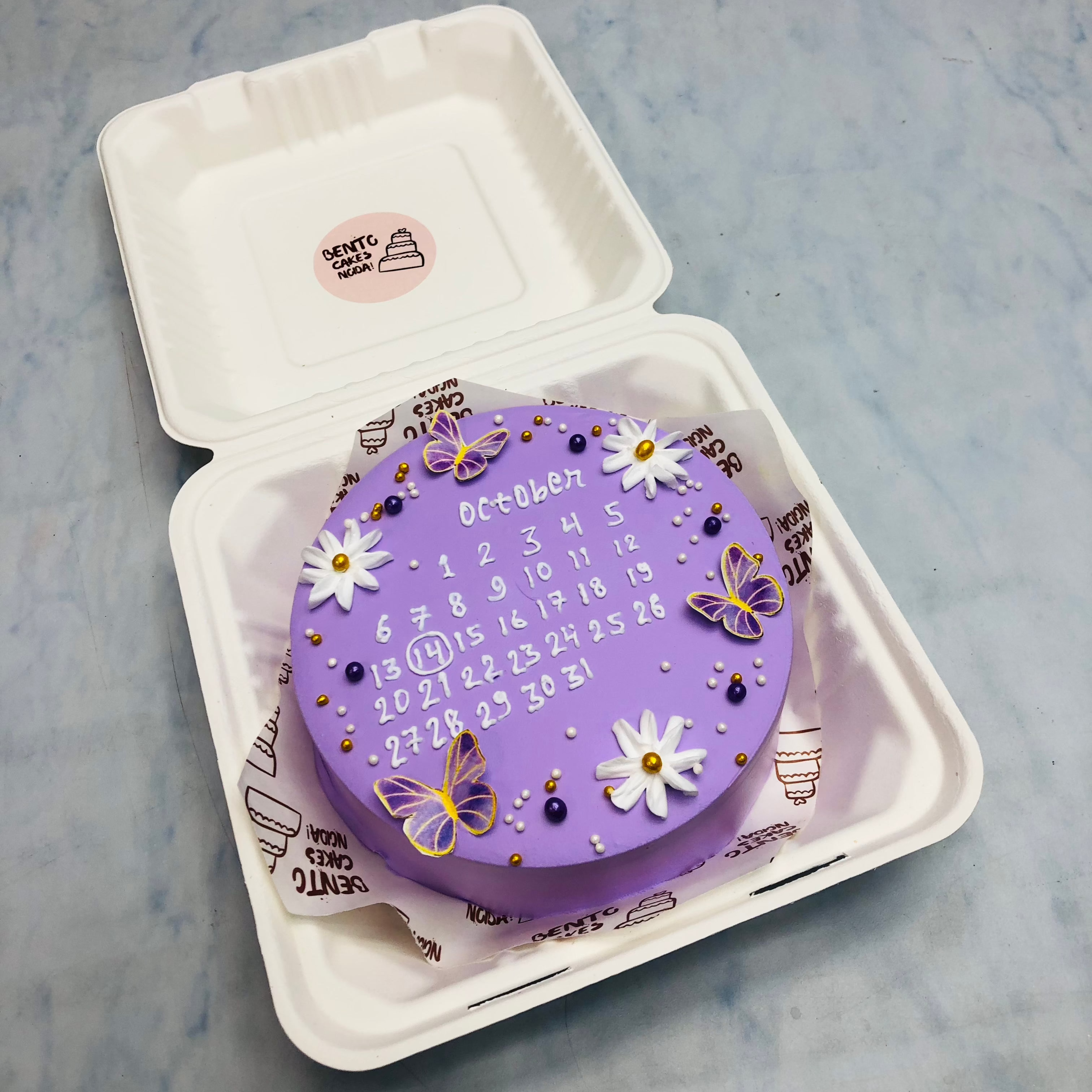 A purple bento cake having pretty floral and butterflies design and a calendar designed on it.