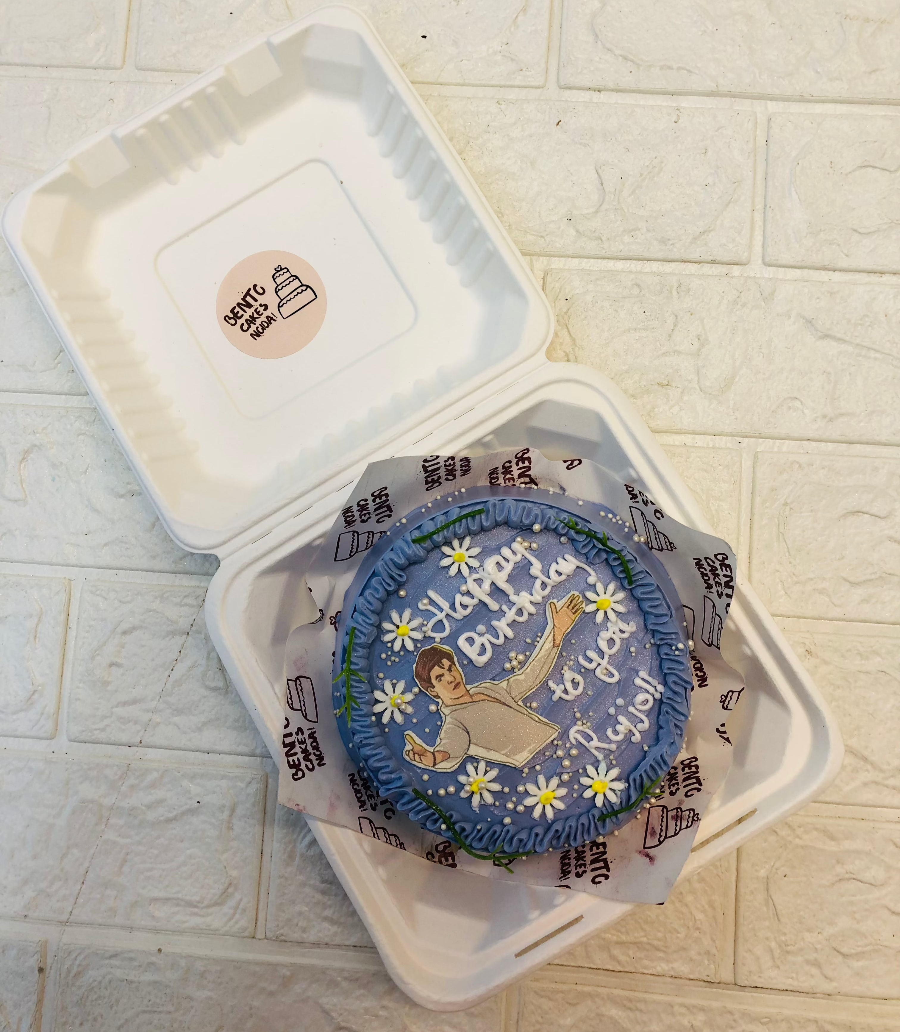 A small round birthday cake with blue icing, decorated with white daisies and an illustration of a person holding a cake. The cake features a personalized message, 'Happy Birthday to you, Rijjo!'