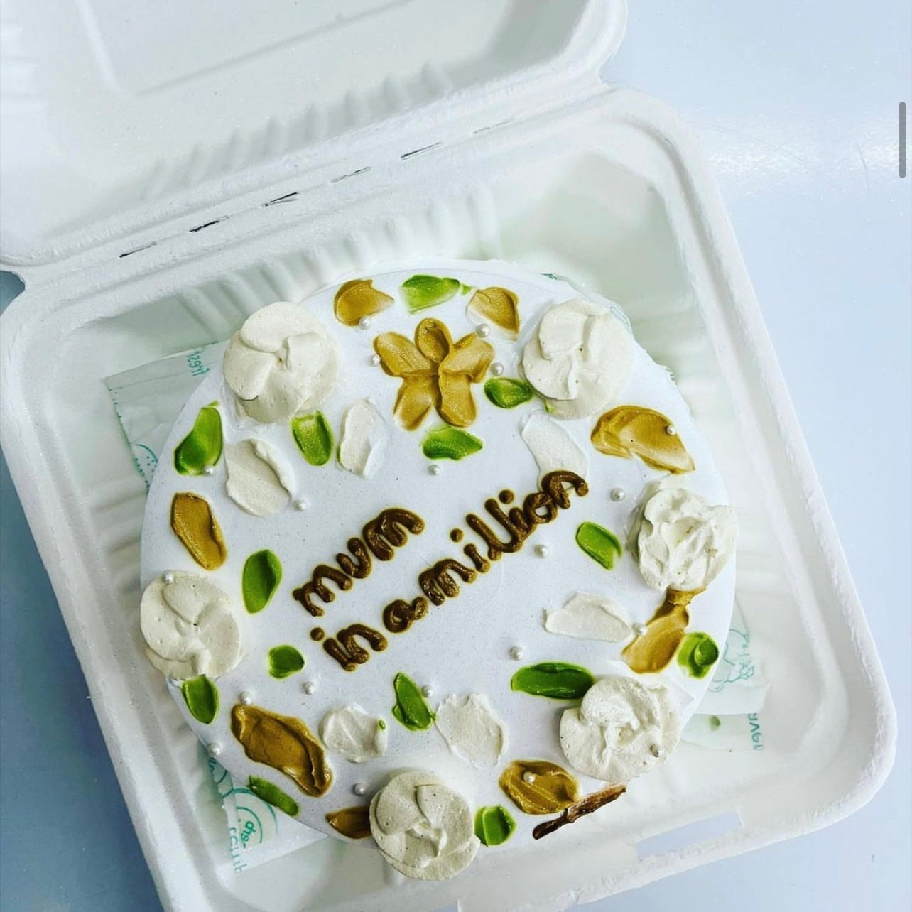 A beautifully decorated round cake with white frosting, adorned with gold and green floral designs, and the words "mum in a million" written in gold icing. The cake is placed inside a white bento box.