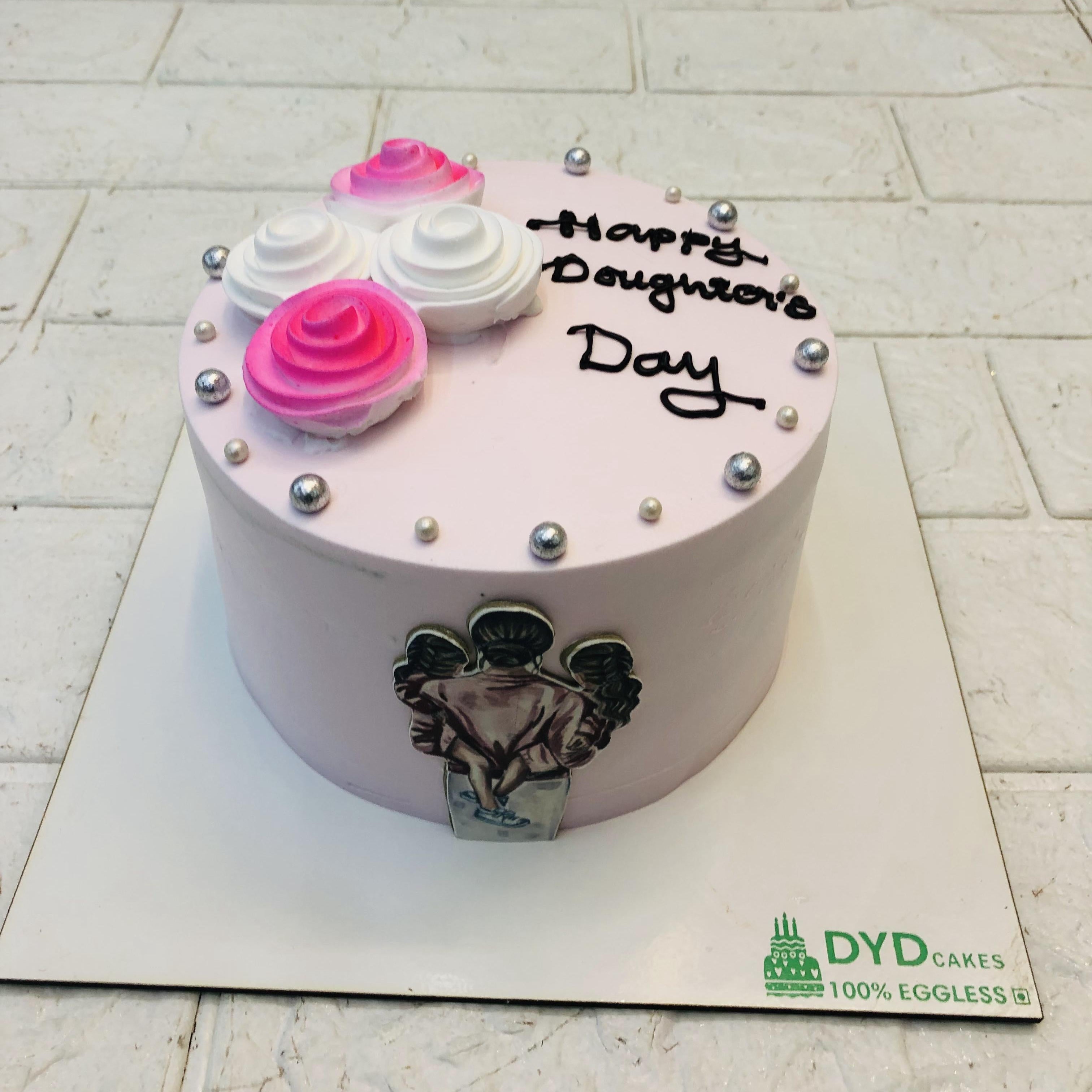 A light pink cake decorated with white and pink creamy floral design and decorated with silver sprinkles with a mother and daughters print on its side.