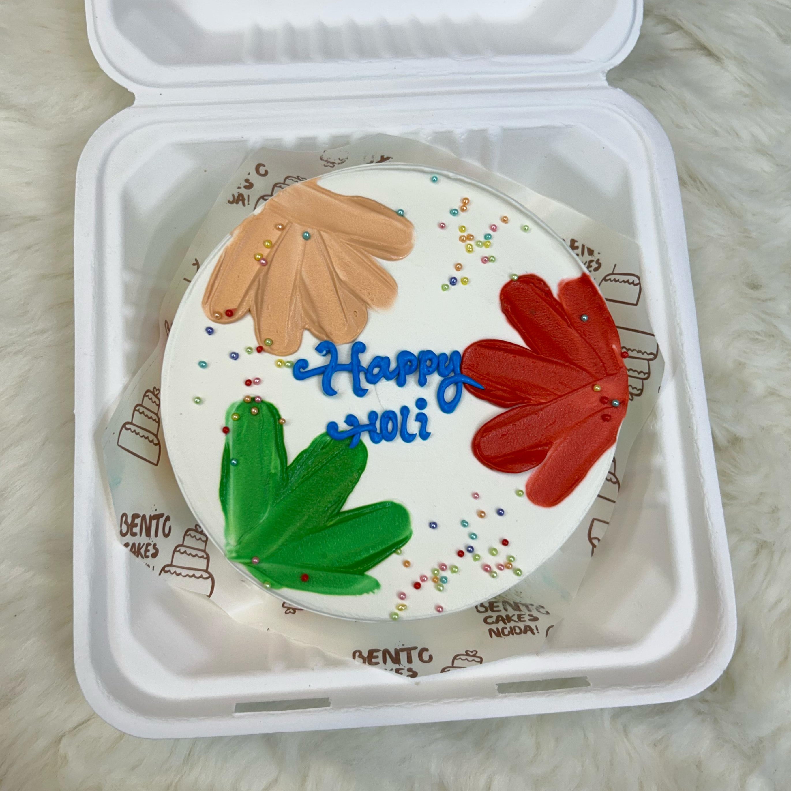 A round cake with colorful decorations and the text "Happy Holi" written on it, placed inside a white takeaway box. The cake has three large flower-like designs in orange, red, and green, with small multicolored sprinkles scattered around with front view.