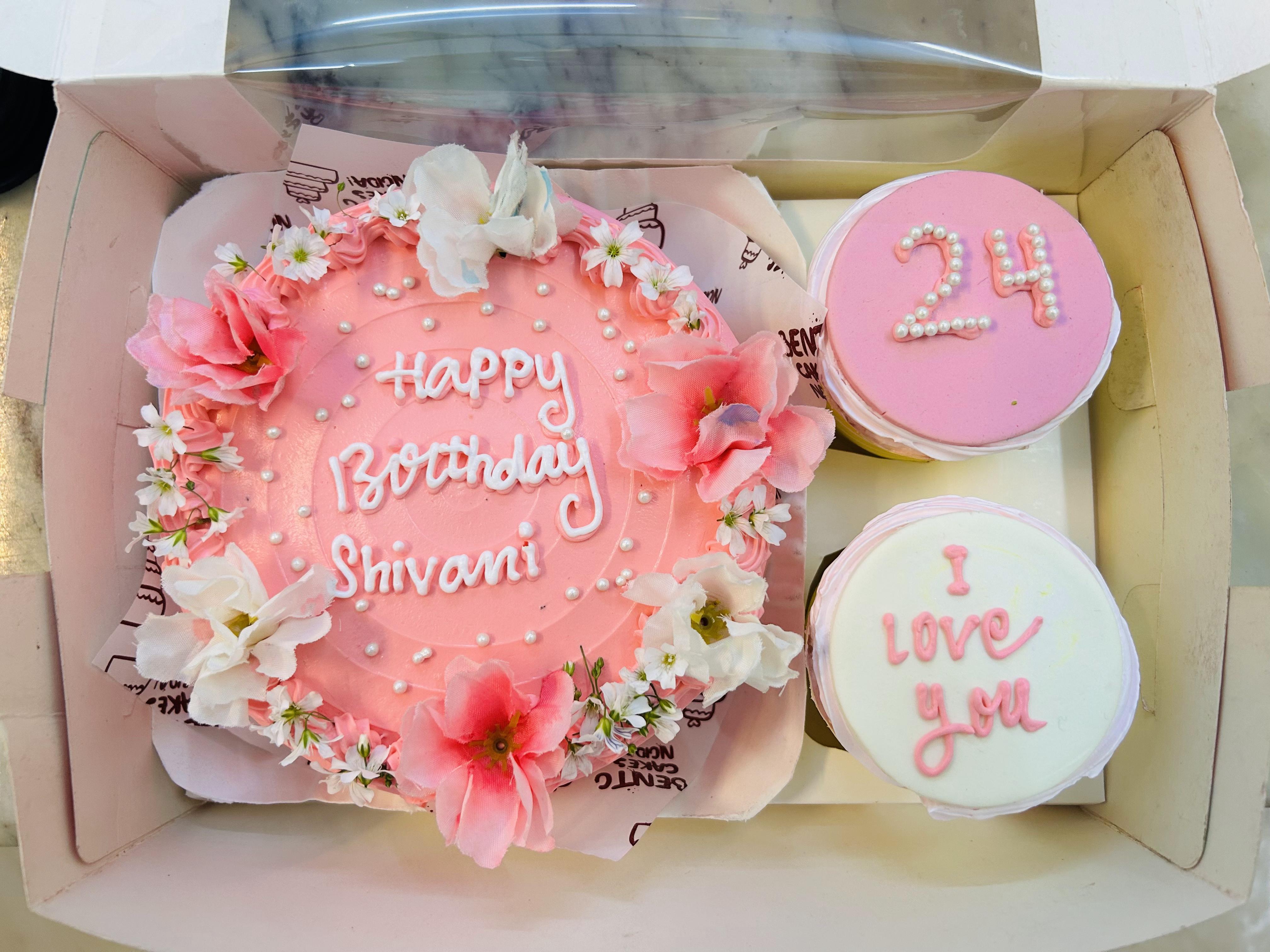 A floral-themed mini bento cake combo featuring a pink round cake decorated with delicate white and pink flowers, small white pearls, and the text "Happy Birthday Shivani" in white icing. Accompanying the cake are two cupcakes: one with pink frosting and the number "24" crafted from pearls, and the other with white frosting and the text "I love you" written in pink icing.