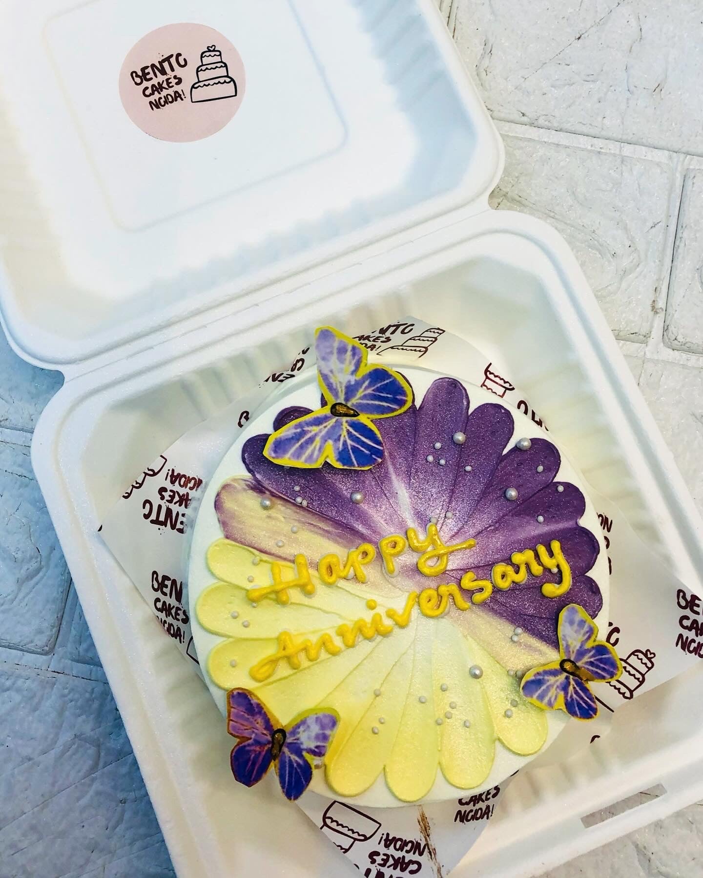 A white bento cake with a yellow and purple floral design for "Happy Anniversay" with blue butterflies.