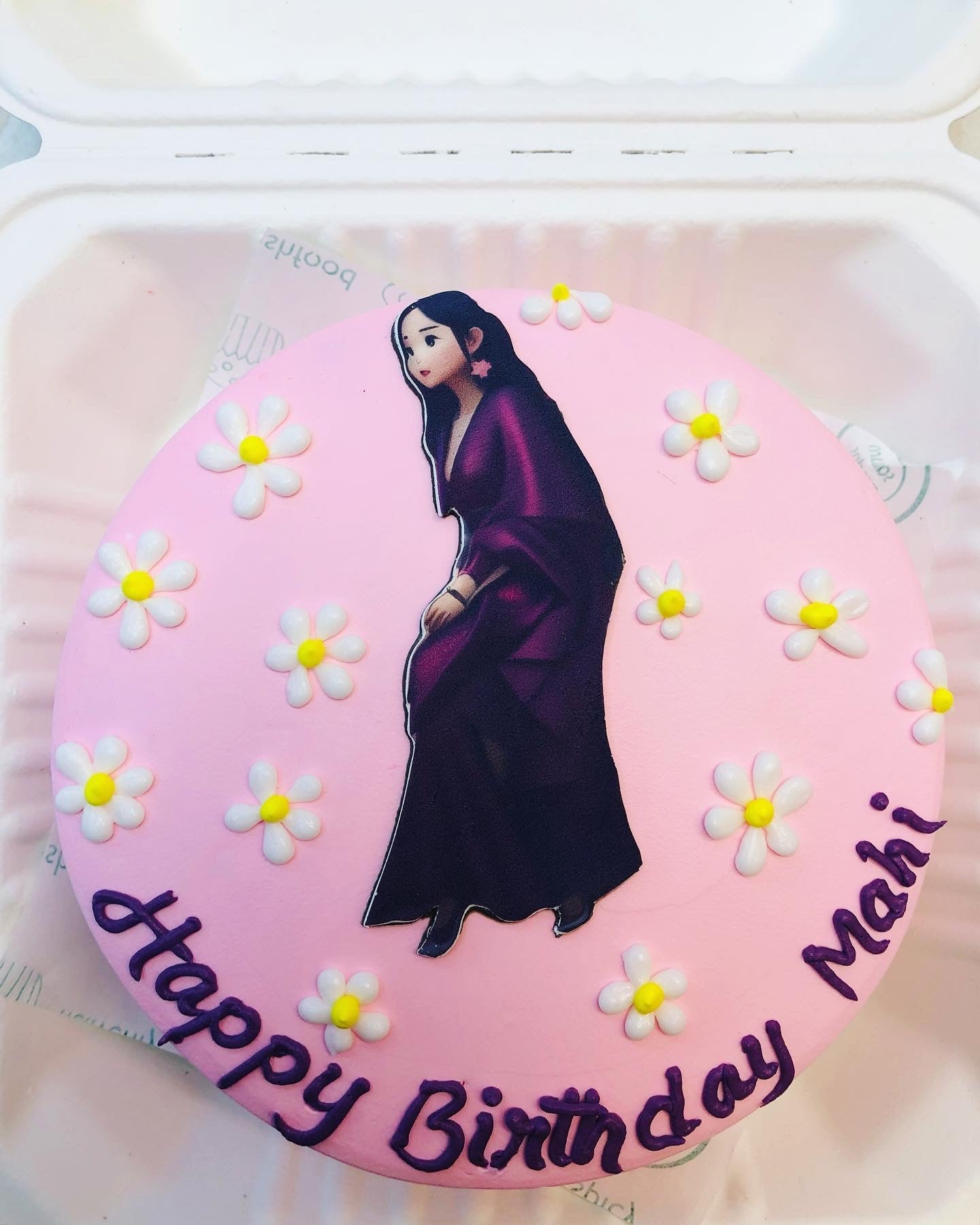 A light pink cake decorated with white floral designs and a prink of girl in purple saree placed on it wit a happy birthday Mahi text.