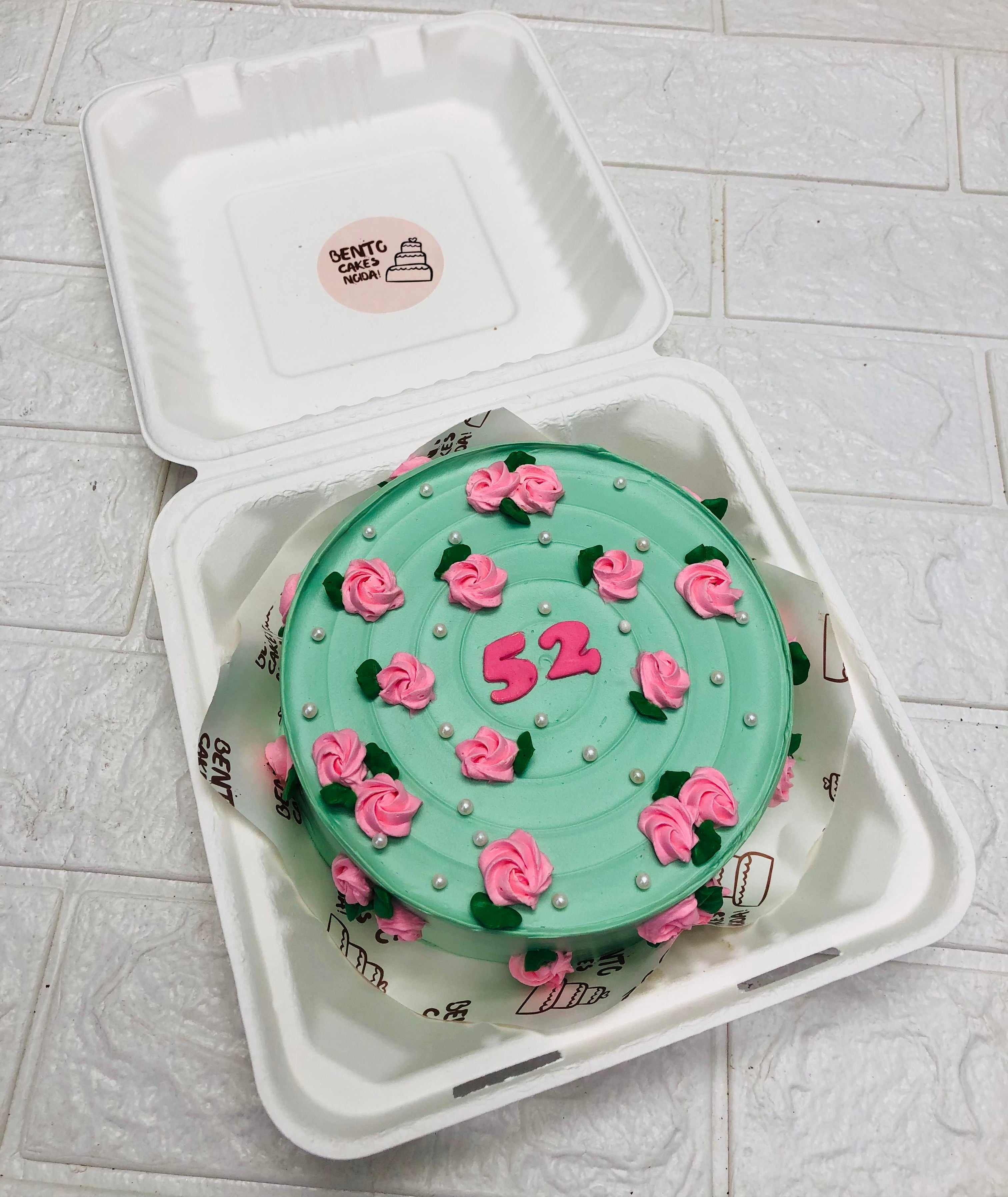A beautifully decorated round birthday cake with soft green frosting, adorned with delicate pink rose-shaped icing and tiny white pearls. The number 52 is crafted in pink icing at the center, making it a perfect cake for a milestone birthday. 