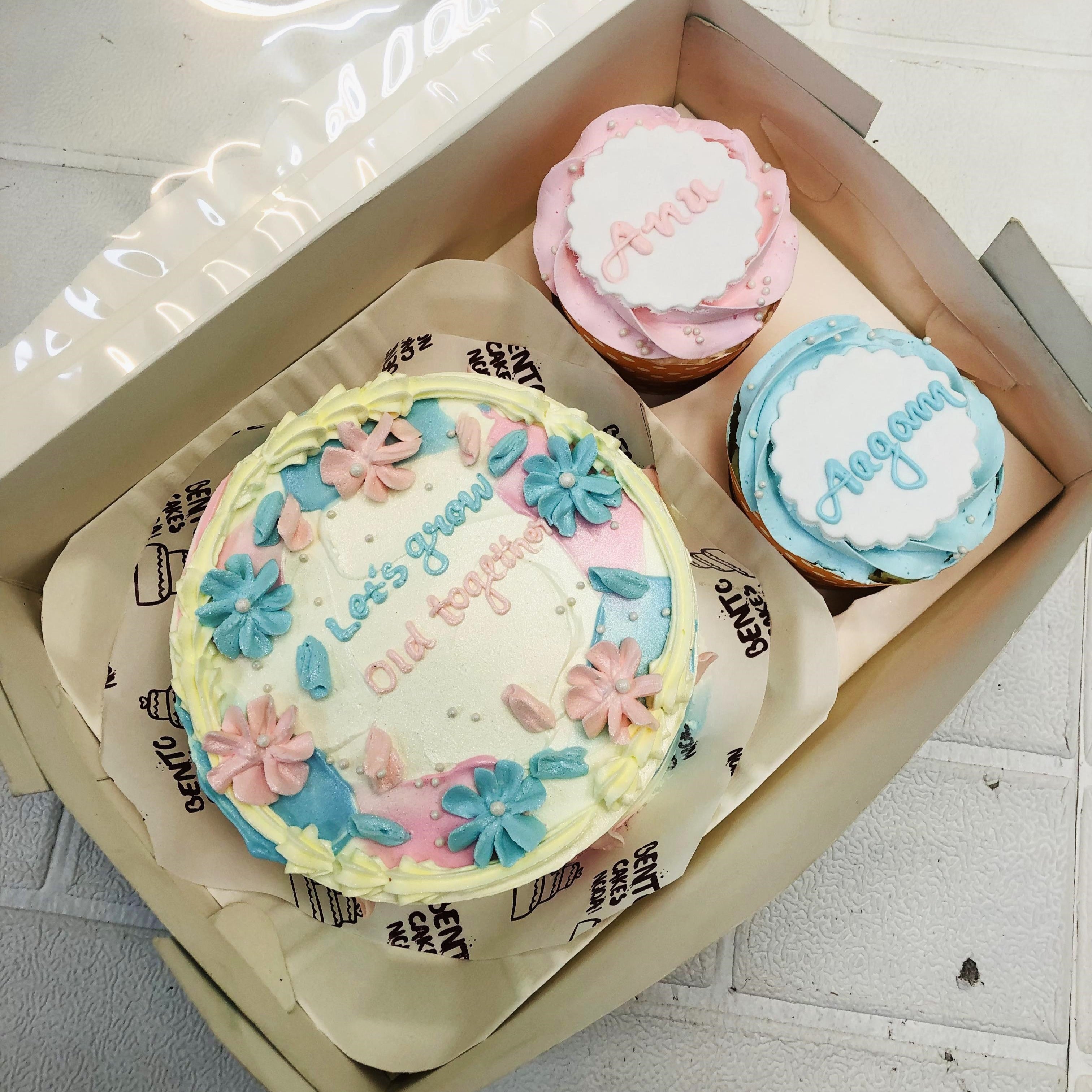 A Floral Theme Mini Bento Combo Cake set featuring a round cake with pastel floral designs and the message "Let's grow old together" in blue and pink icing. Accompanied by two cupcakes, one with "Anni" in pink icing and the other with "Aagam" in blue icing.