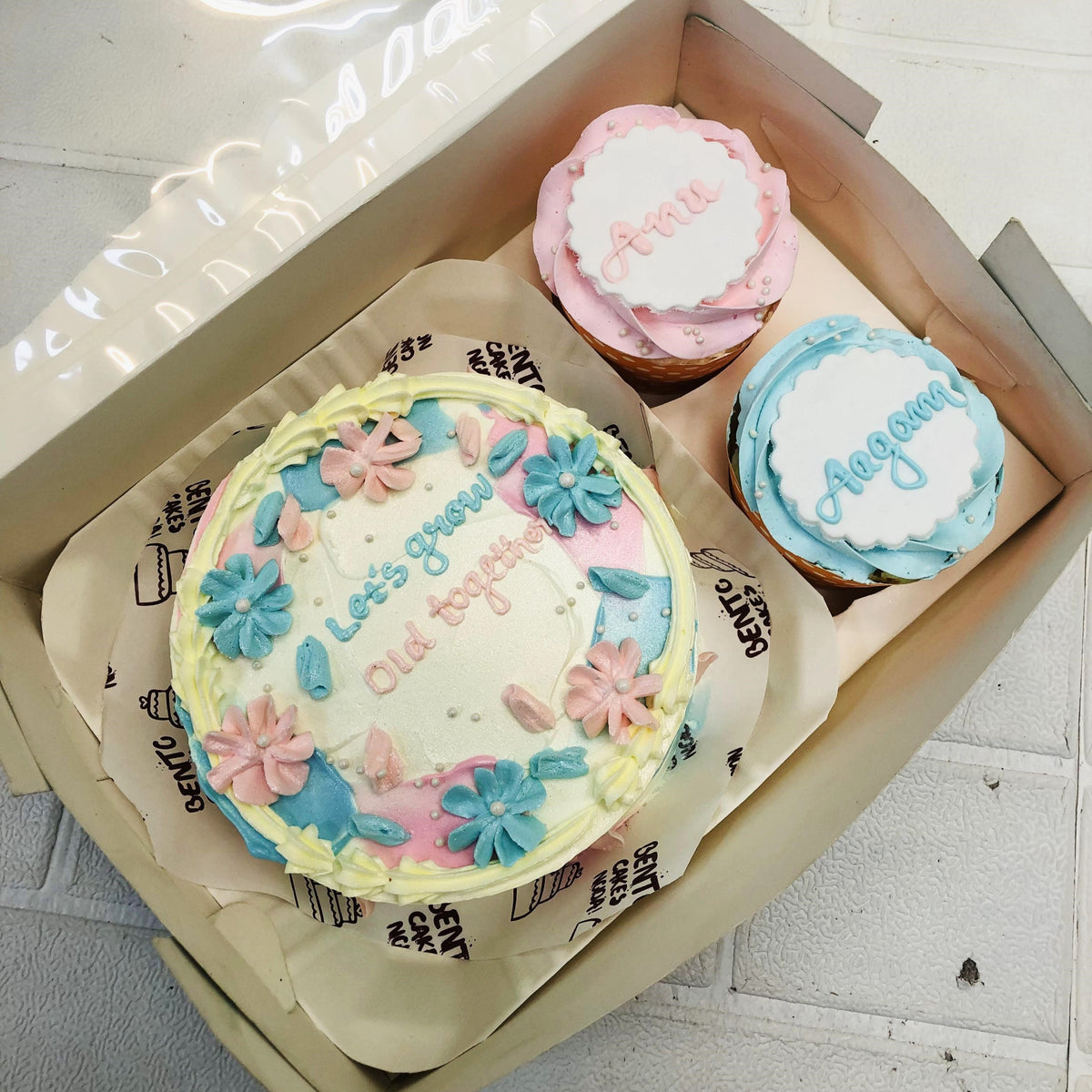A Floral Theme Mini Bento Combo Cake set featuring a round cake with pastel floral designs and the message "Let's grow old together" in blue and pink icing. Accompanied by two cupcakes, one with "Anni" in pink icing and the other with "Aagam" in blue icing.