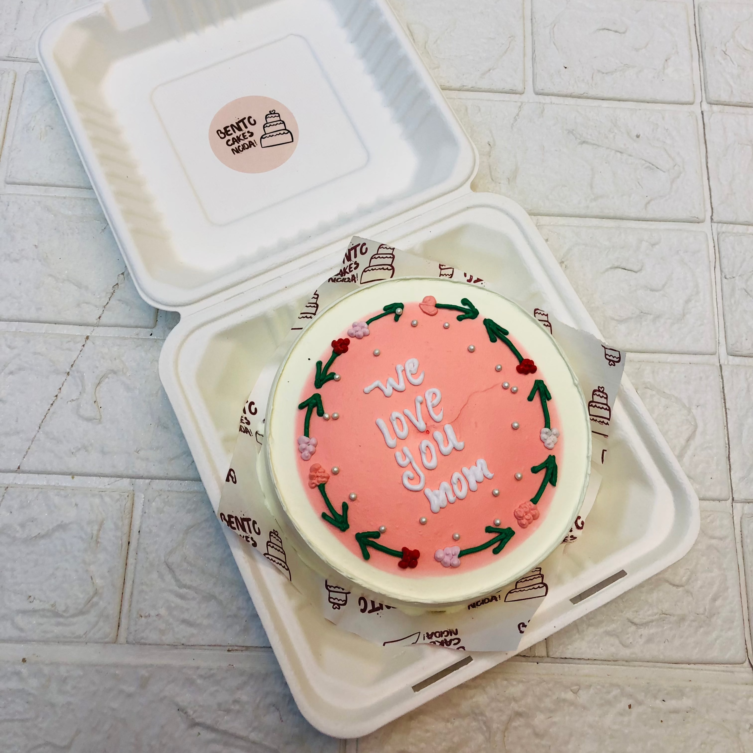 A white cake designed with pink shade in center and surrounded with floral design with text "We love you mom".