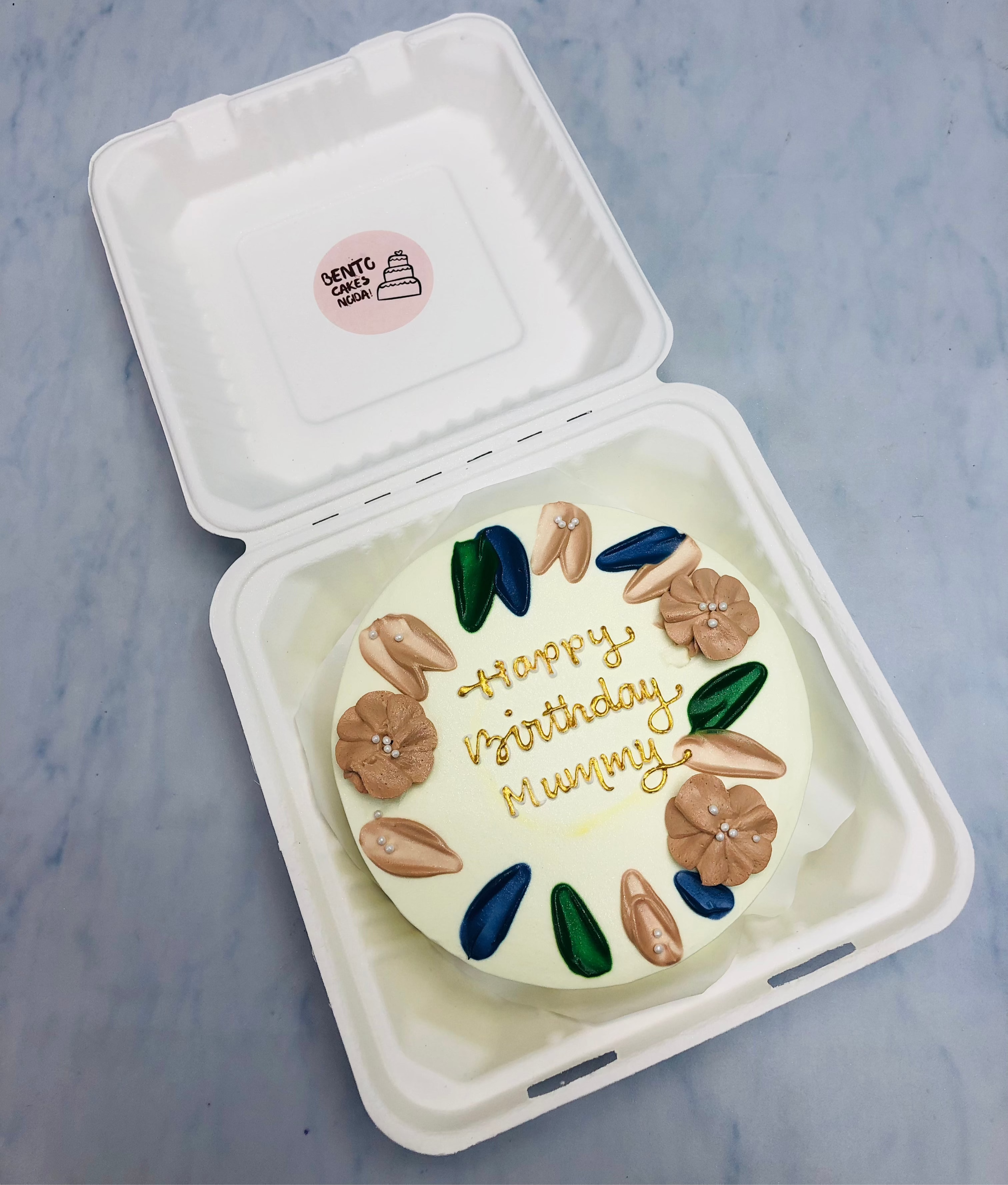 A white bento cake with colorful floral petals design in round shape with a "Happy Birthday Mummy" text written with golden icing.