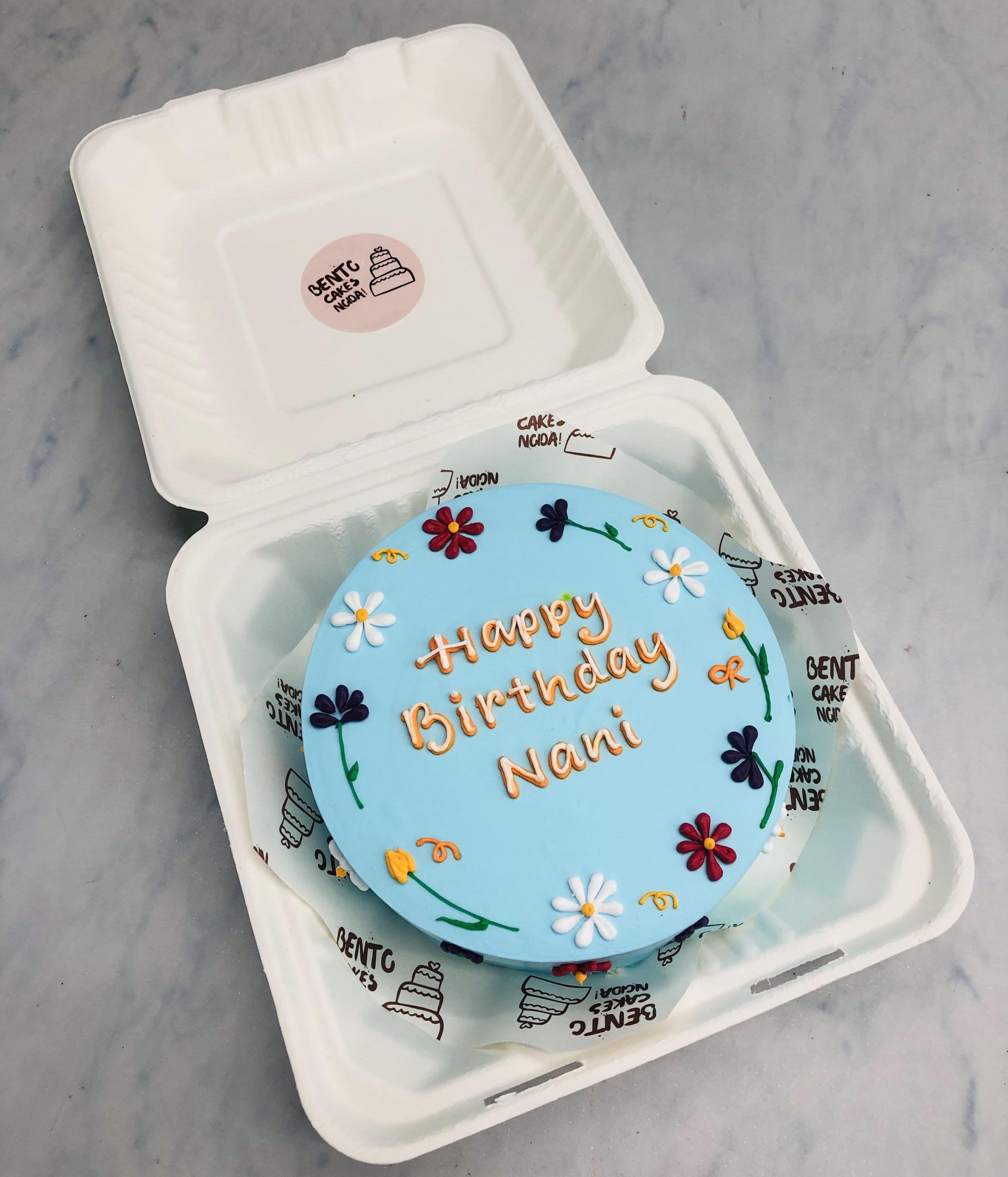 A charming blue birthday cake featuring 'Happy Birthday Nani' in gold icing, adorned with colorful floral decorations, placed inside a bento box.