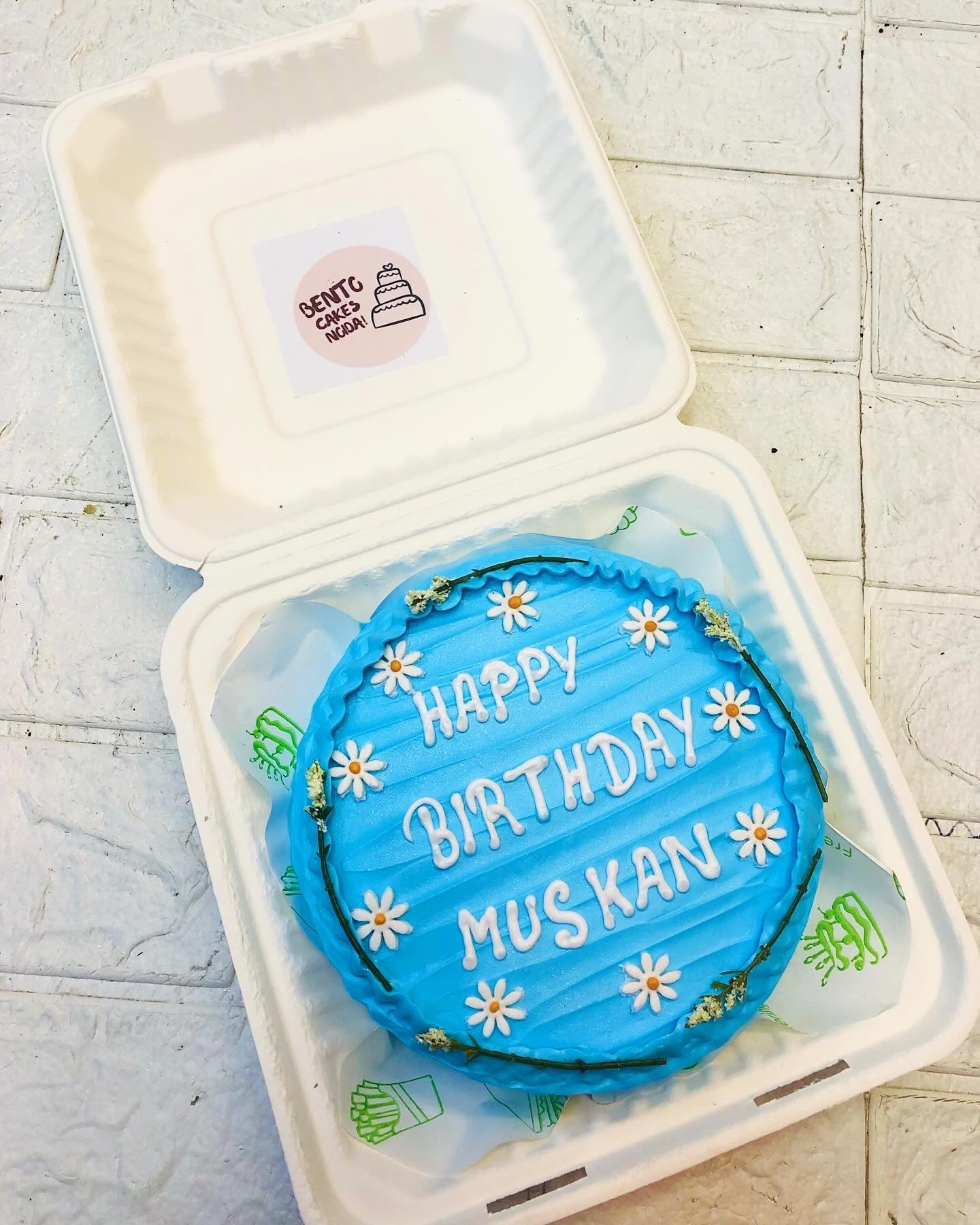 A beautifully crafted blue birthday cake featuring white daisy decorations and the message "HAPPY BIRTHDAY MUSKAN" in delicate white icing. 