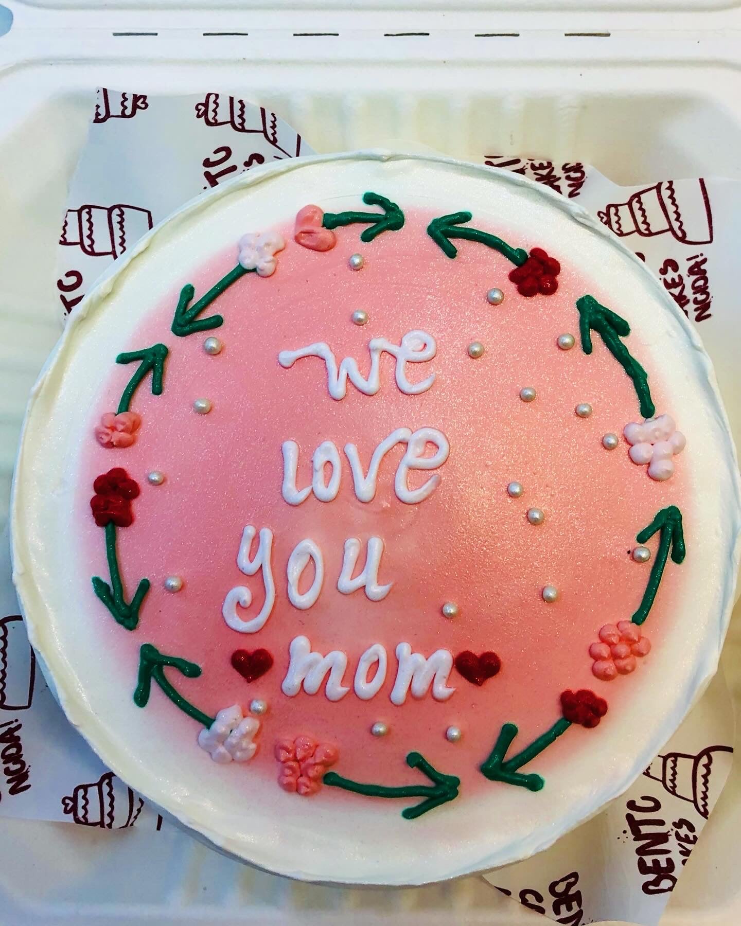 A white cake designed with pink shade in center and surrounded with floral design with text "We love you mom".