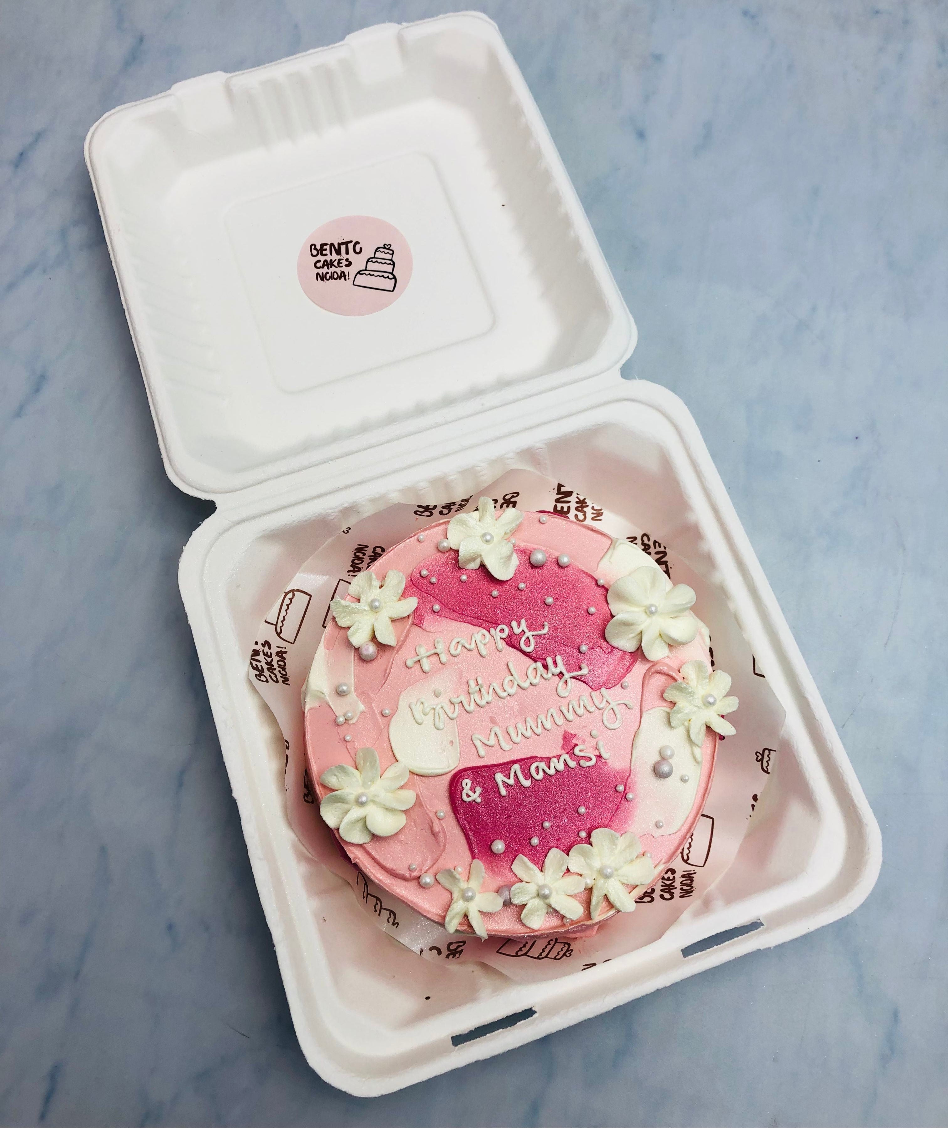 A beautifully decorated pink birthday cake with white floral accents and the message 'Happy Birthday Mummy & Mama' written on it.