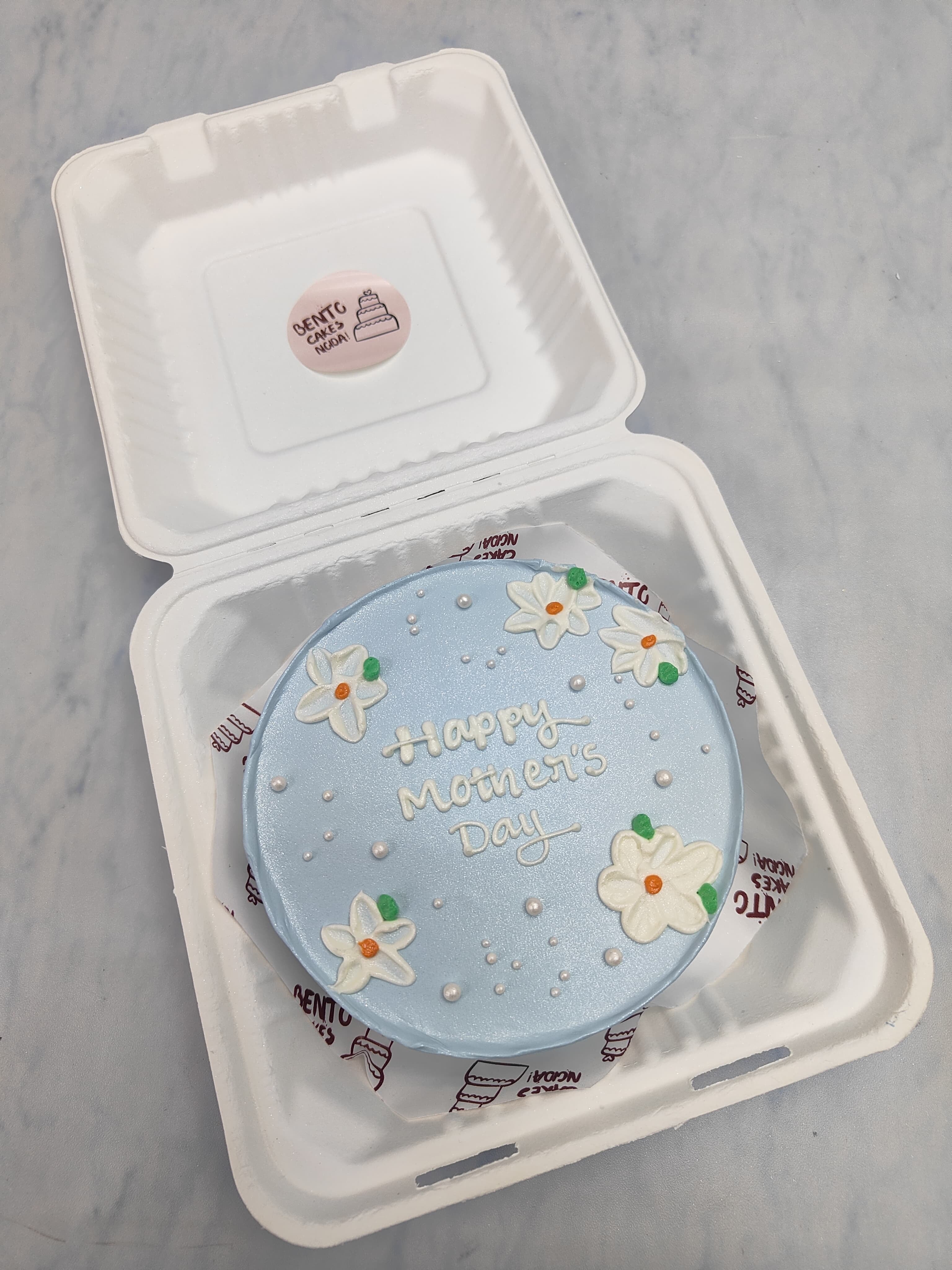 A shiney blue bento cake is designed with white florals on cake top for Mothers Day.
