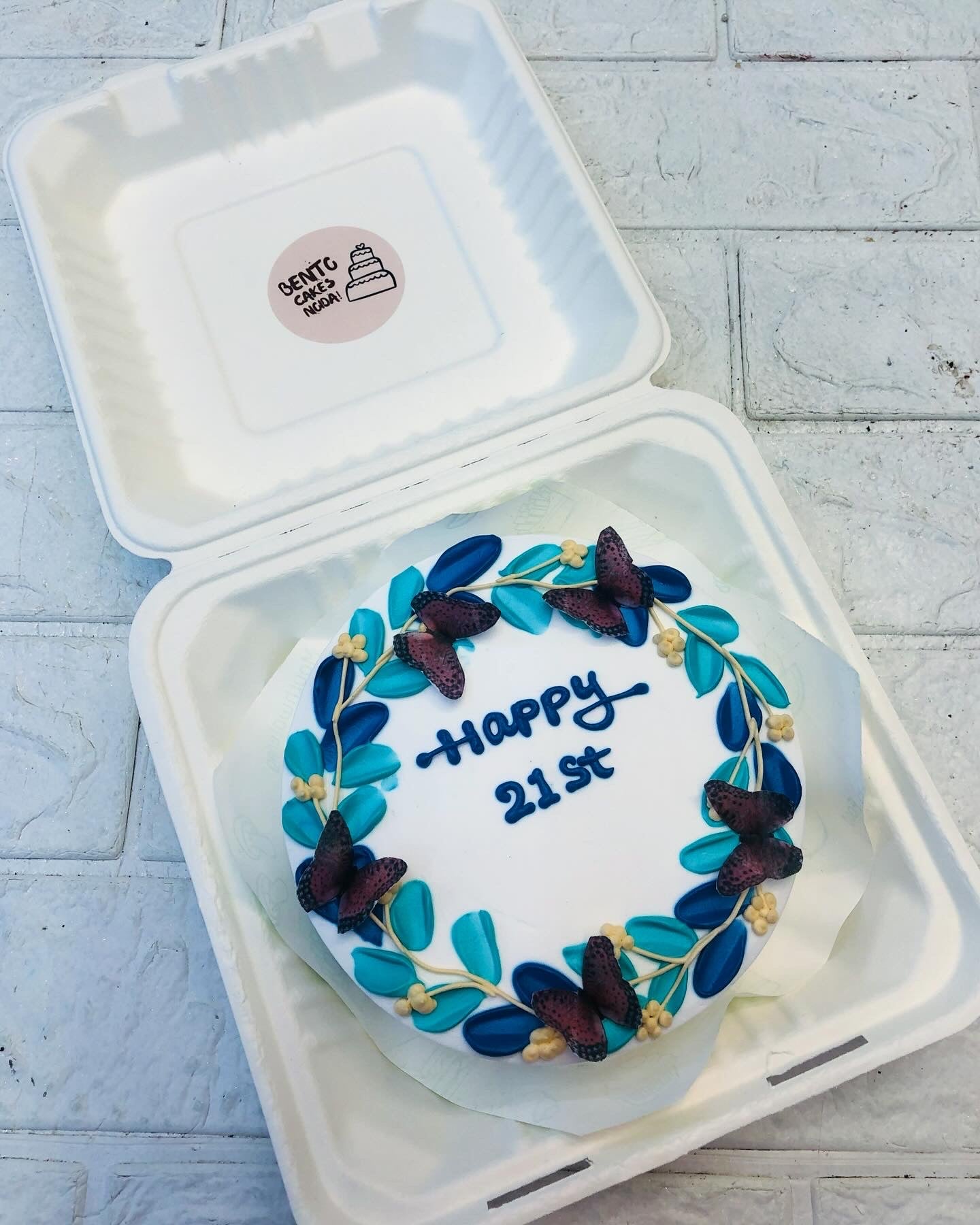 A beautifully decorated white cake inside a box, adorned with blue and purple butterflies, leaves, and floral accents. The words "HAPPY 21st" are written in blue icing in the center.