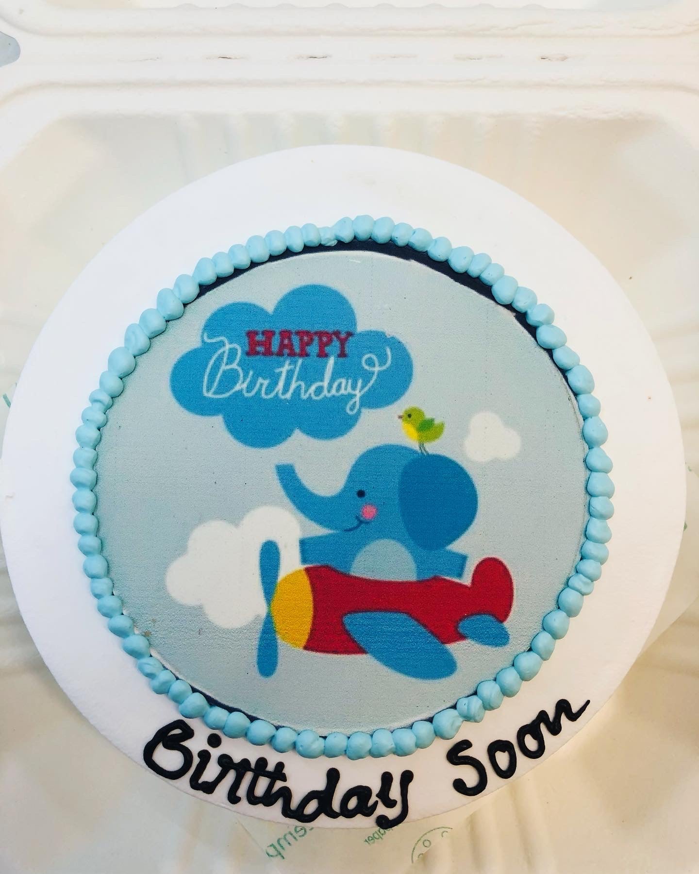 A light blue bento cake is decorated with an elephant flying on plane and Happy Birthday text o  it.