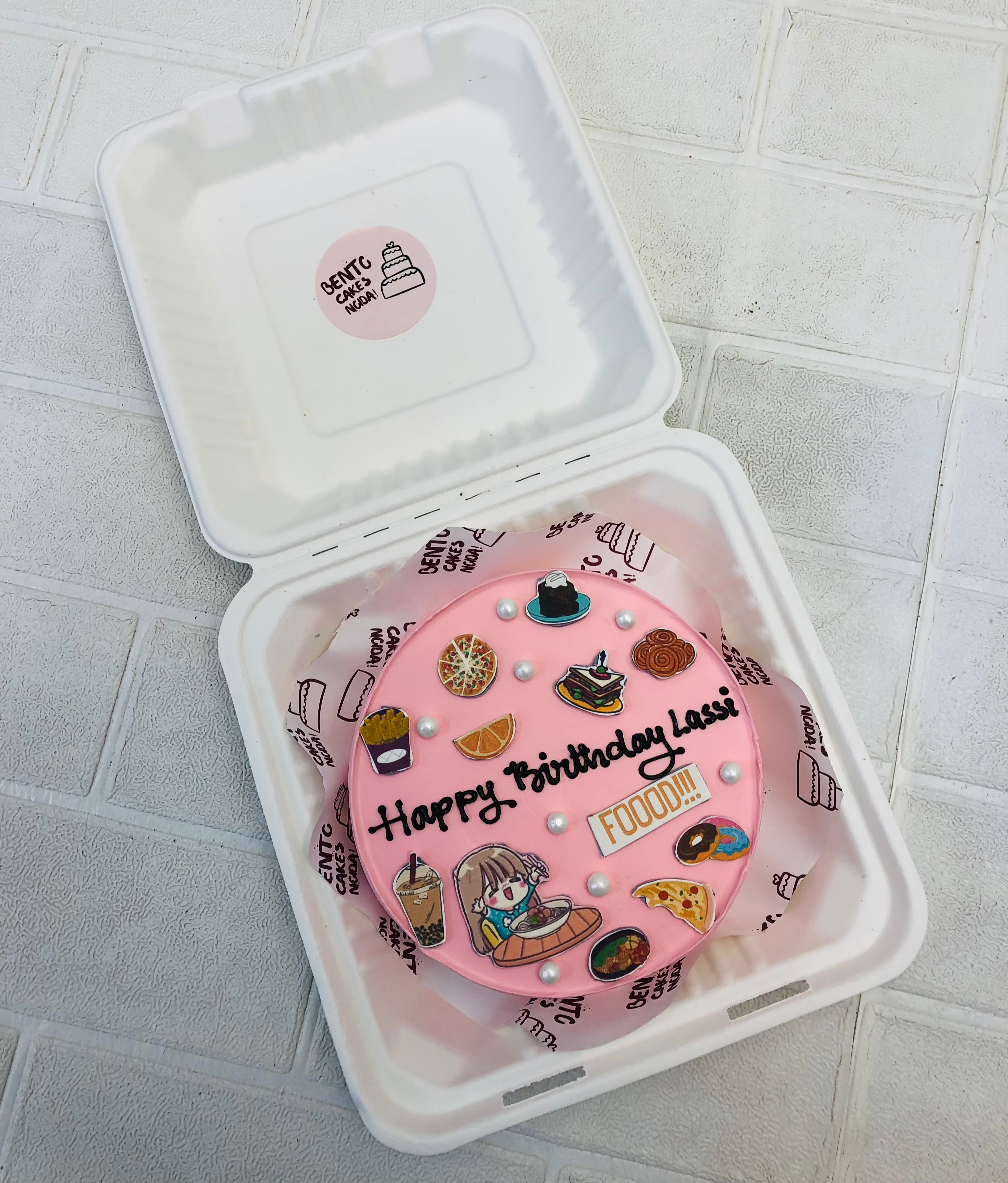 A light pink bento cake is designed for a foody girls birthday whwre all her favourite food items printed on it.