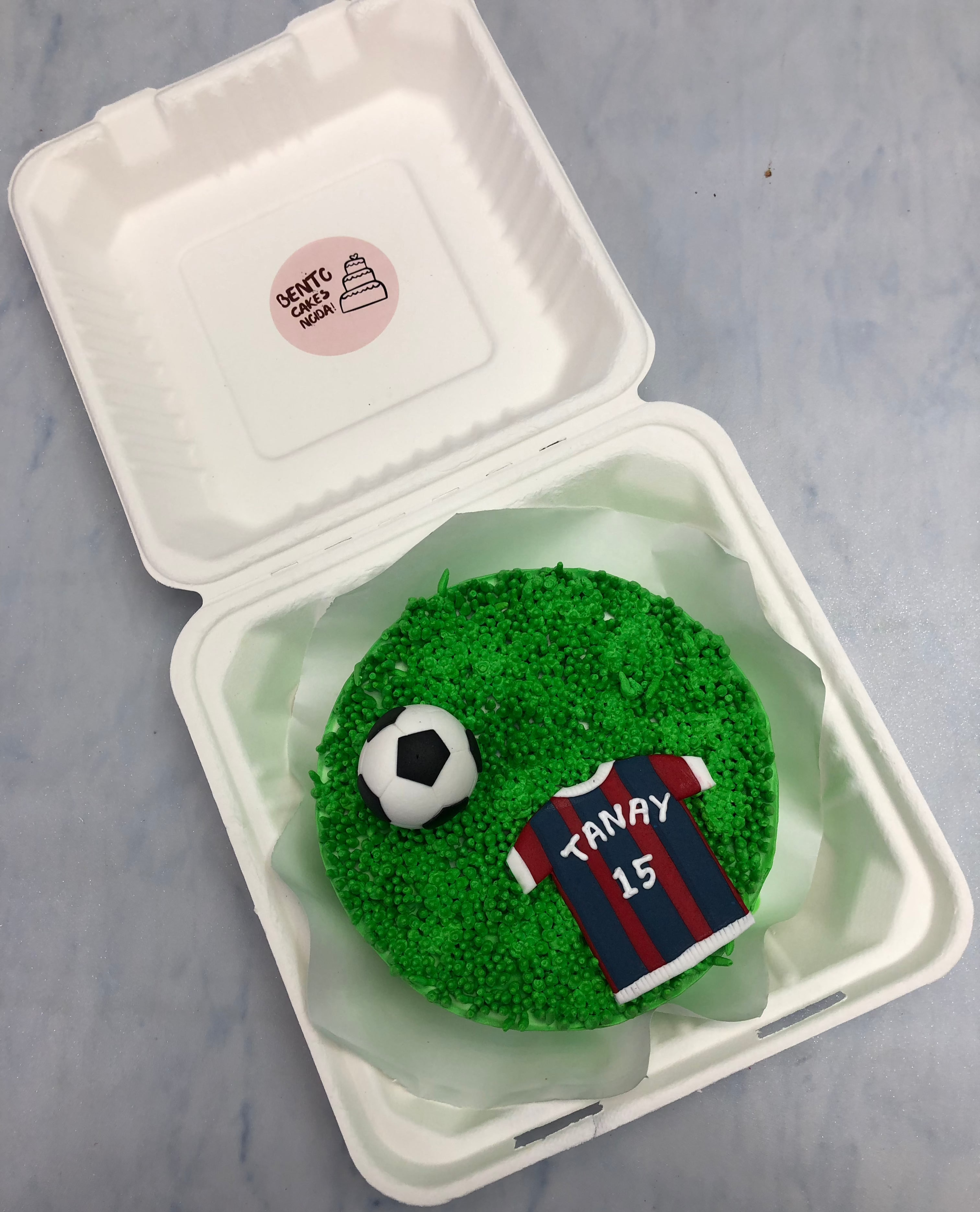 A green bento cake designed on football ground theme and a fondant ball with player dress placed on it.