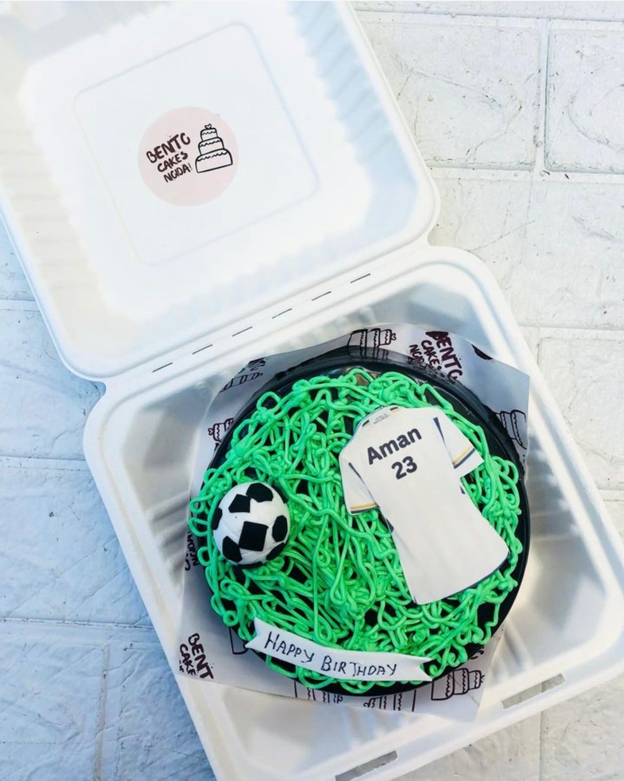 A chocolate bento cake with fondant football design and player jersey and the cake decorated with green cream as football ground theme.