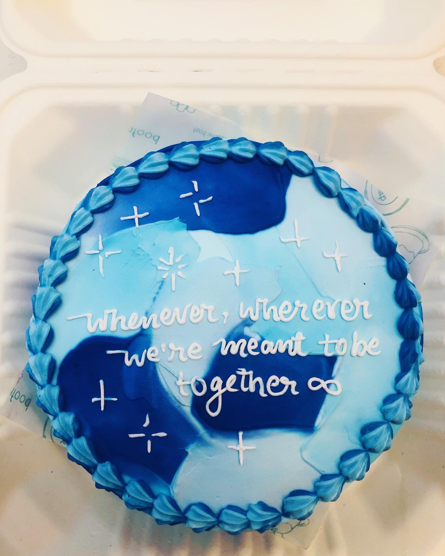 A round cake with blue and white frosting, decorated with small white stars and the message "Whenever, wherever we're meant to be together ∞" written in white icing.