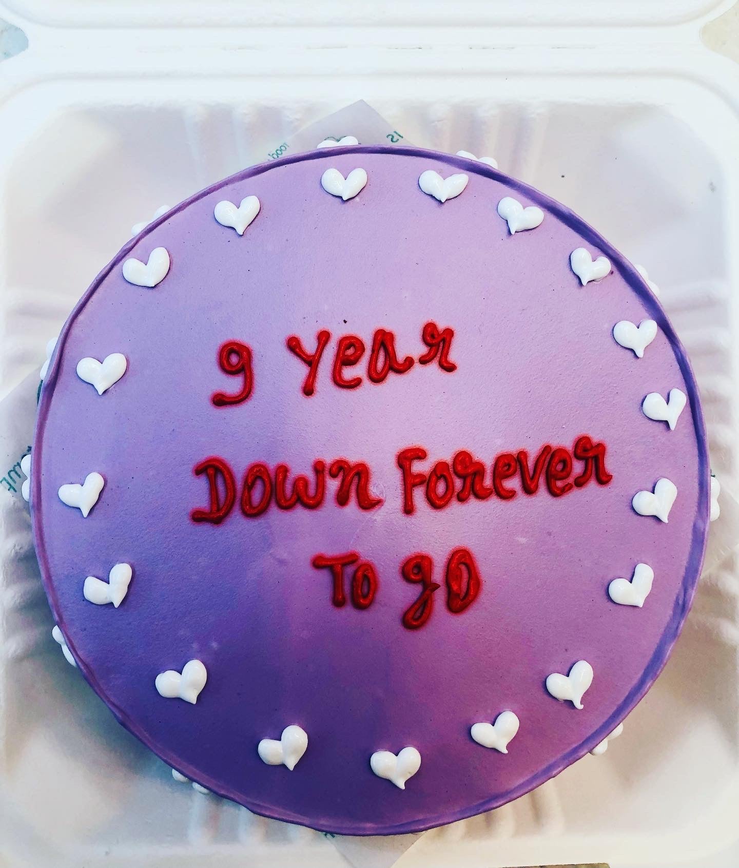 A purple color cake covered with white hearts design on top ands side and a text written with red icing on it. 