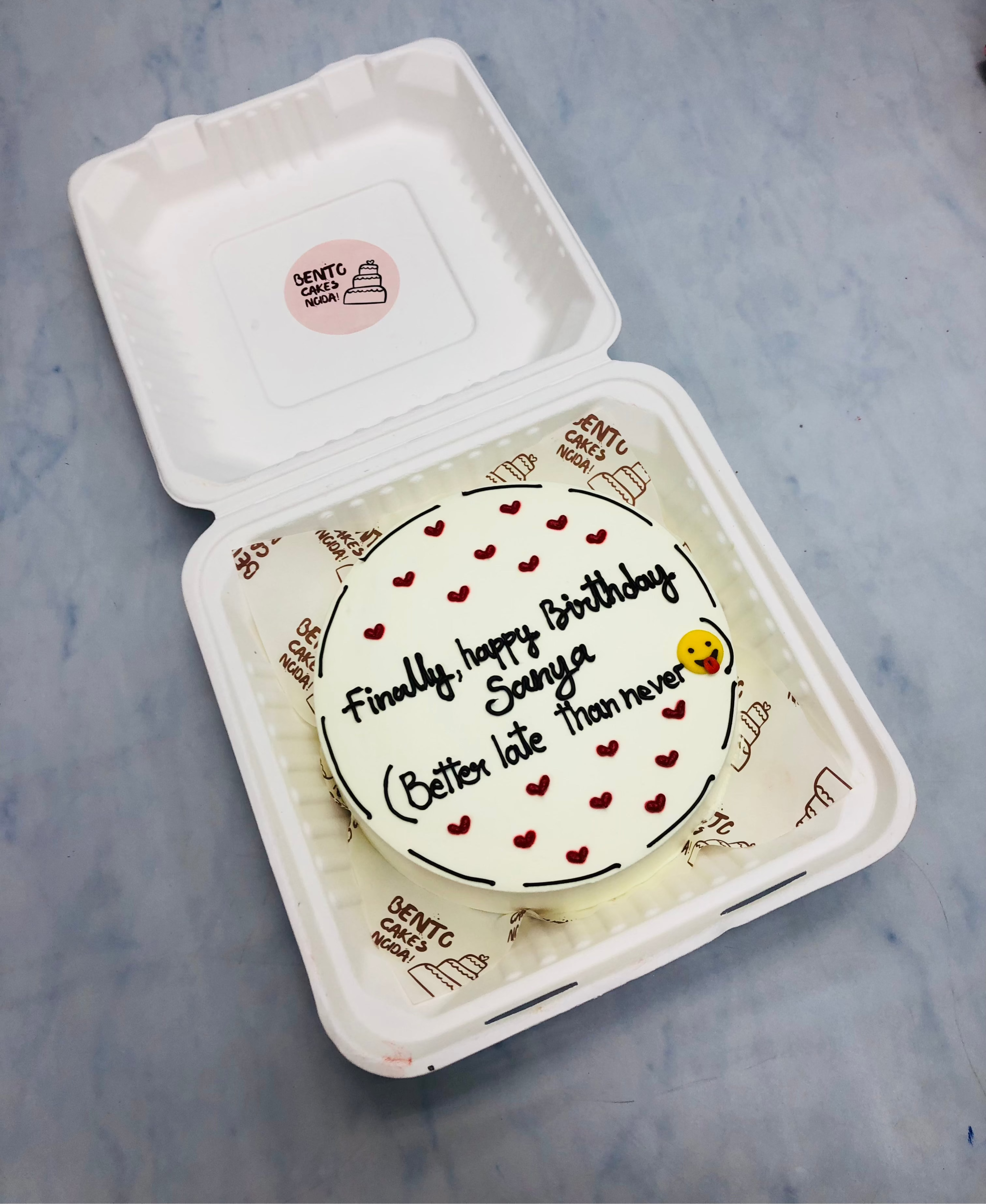 A bento cake with white icing decorated with little red hearts and emoji design.