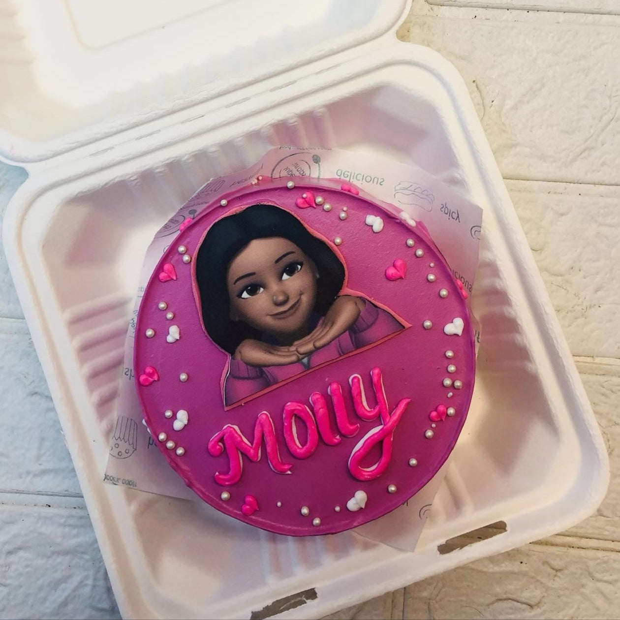 A dark pink cake is decorated with a cartoon cha acter girl on it and a text of "Molly" written with pink icing on it. 