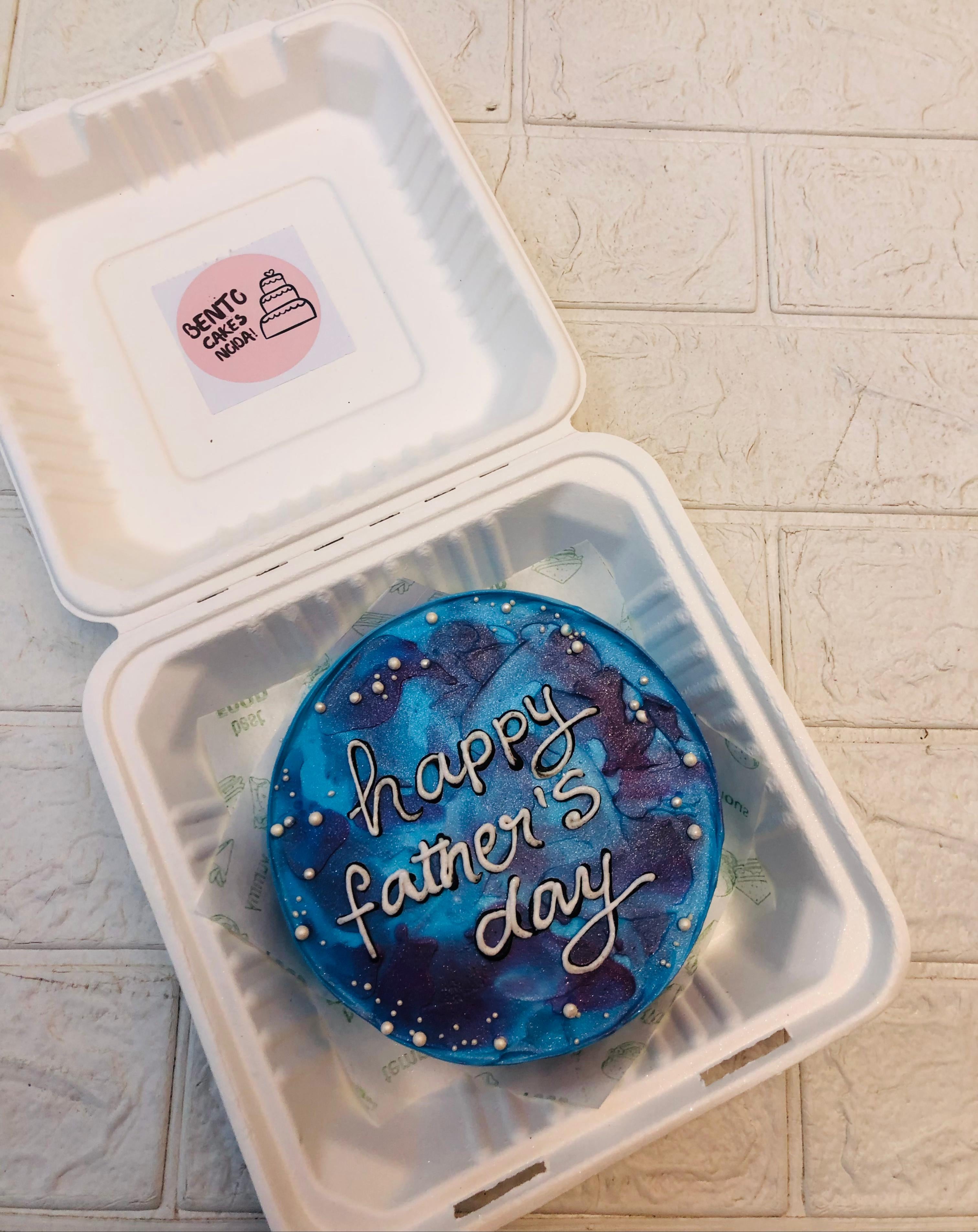 A galaxy-themed bento cake with "Happy Father's Day" written in white icing, placed inside a white bento box with a label reading "Bento Cake Macar" featuring a simple cake illustration.