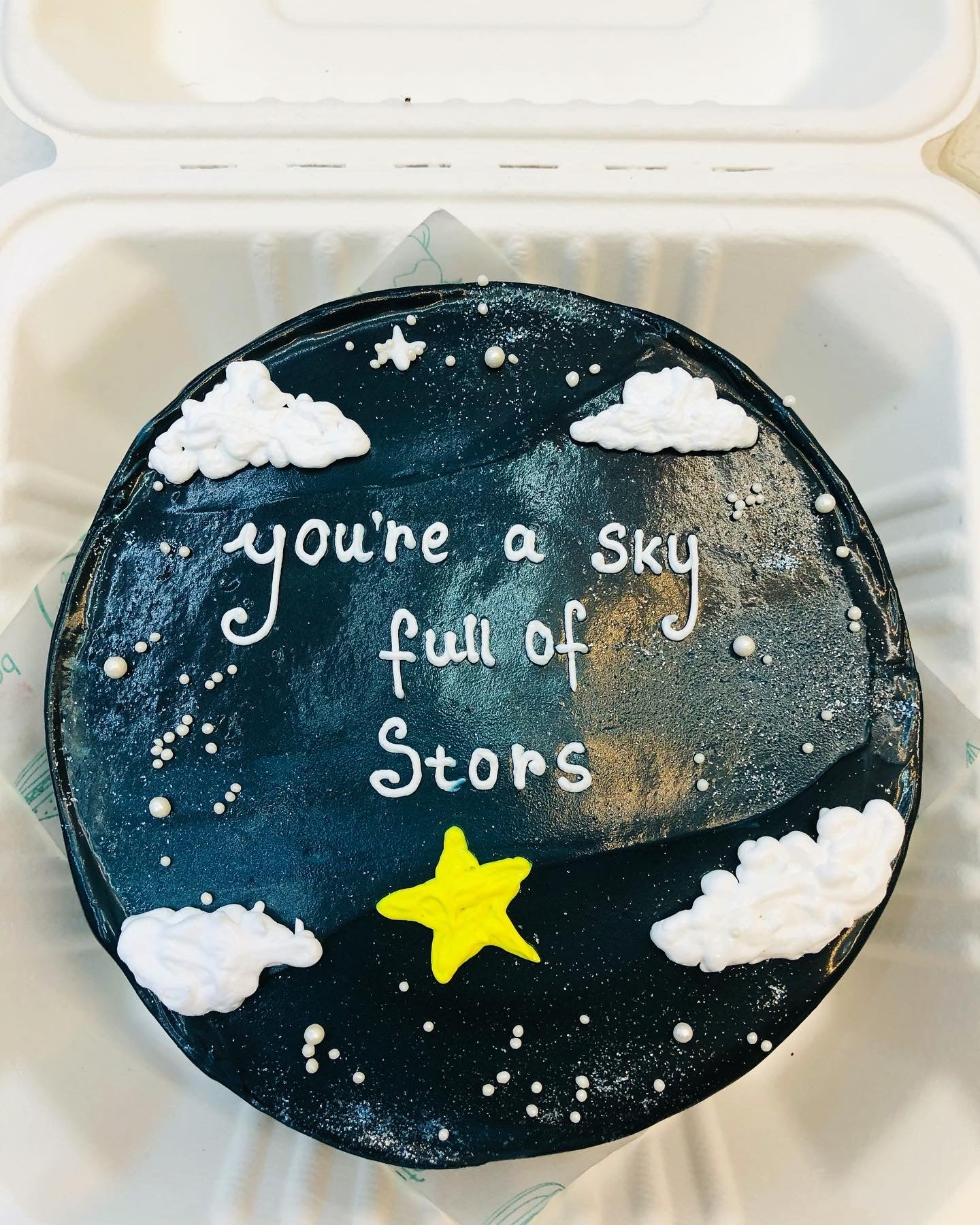 A round cake with a black frosting decorated with white clouds, small white dots resembling stars, and a yellow star. The cake has the text "you're a sky full of stars" written on it.