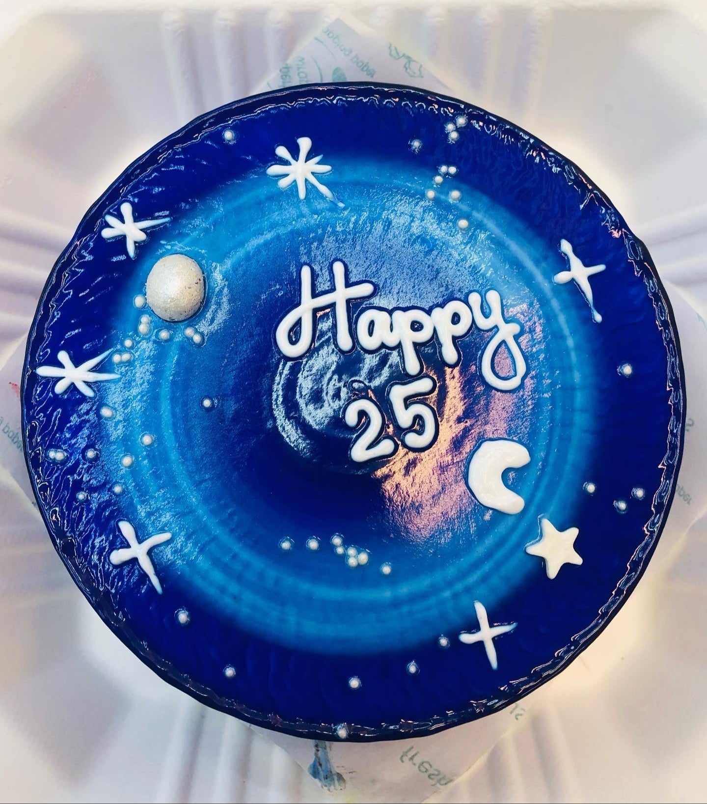 A round cake with a blue galaxy theme, decorated with white stars, a crescent moon, and the text "Happy 25" written in white icing.