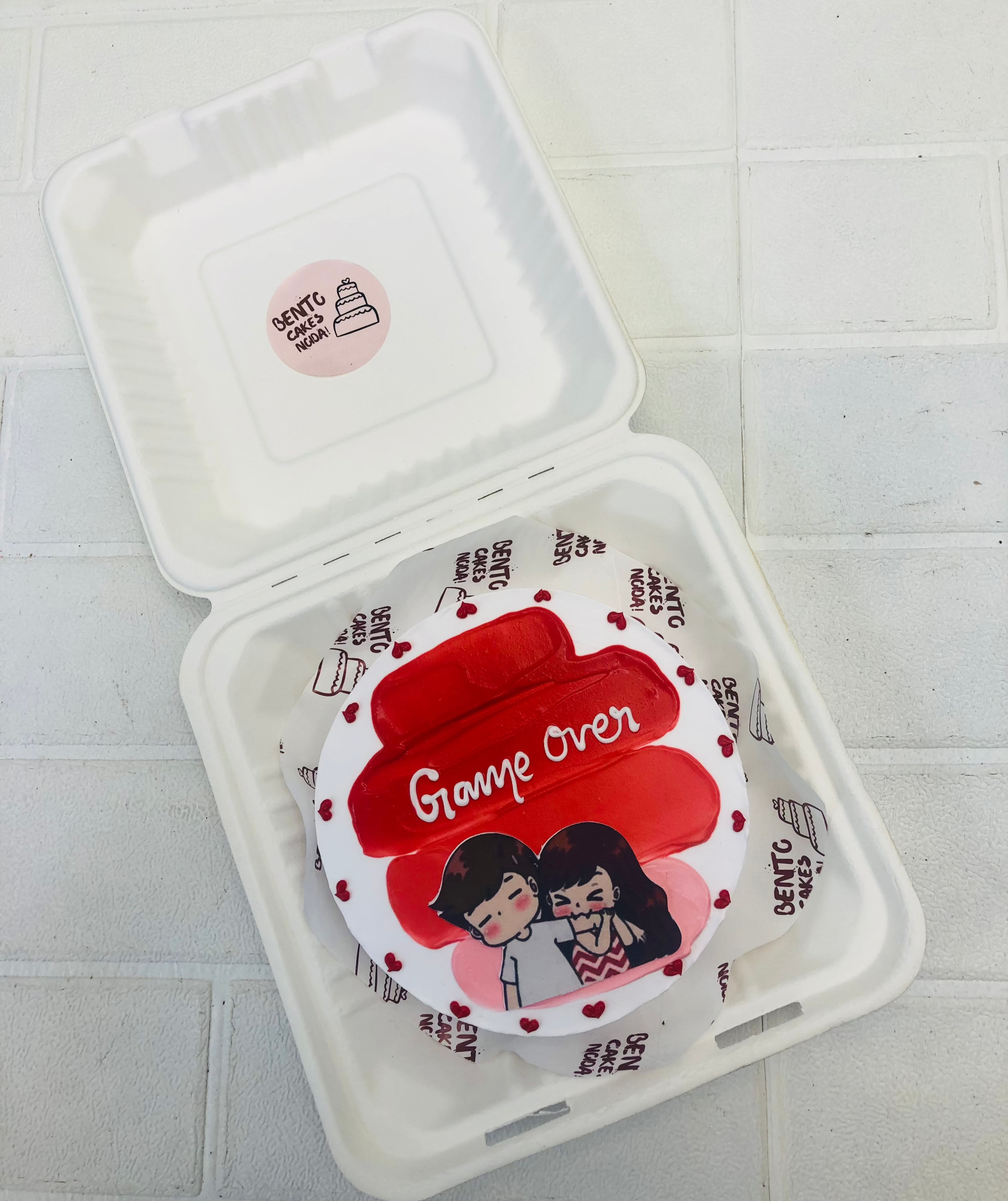 A white bento cake with red shades in center with a biting couple print and decorated withlittle red hearts.