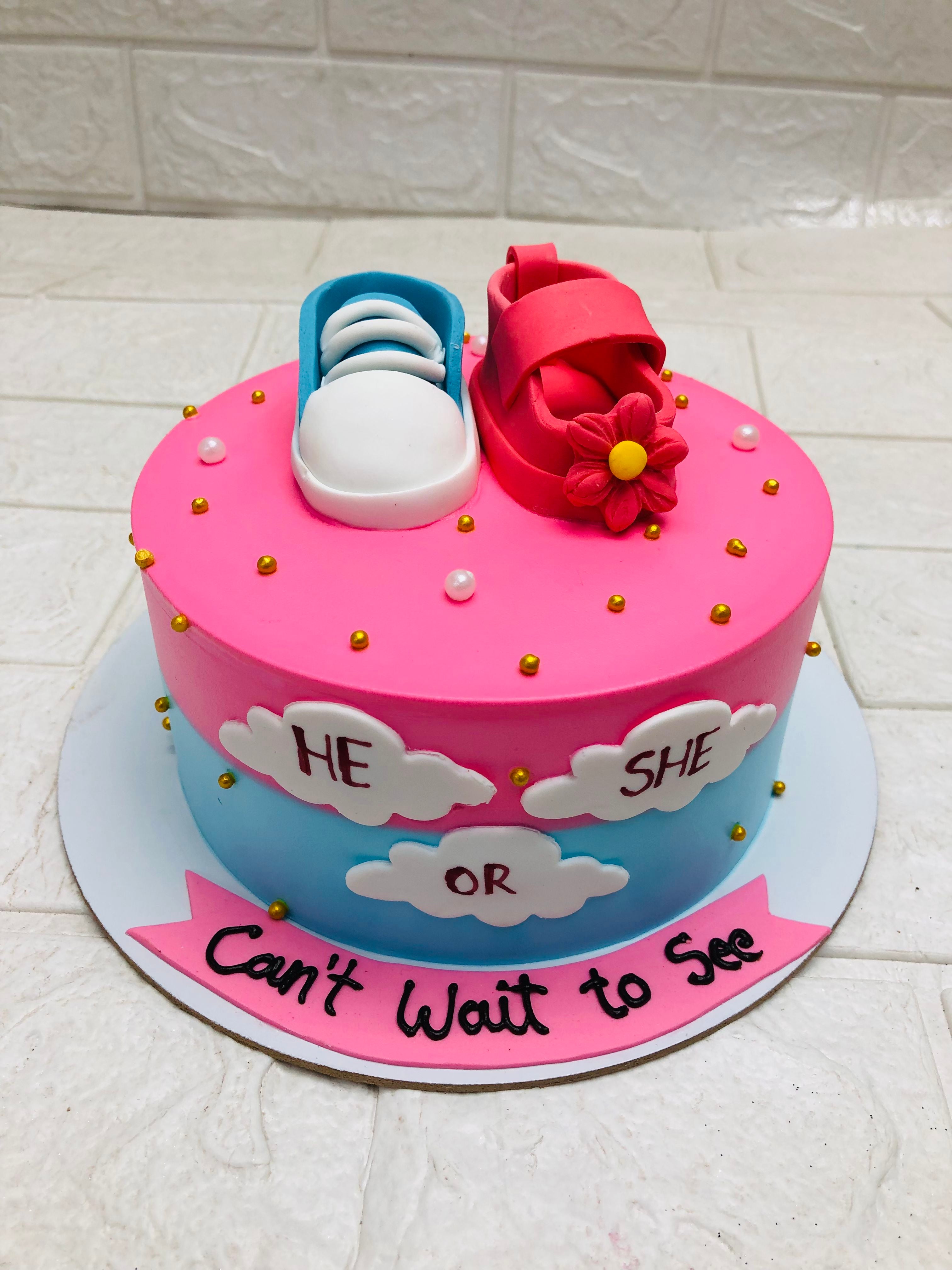 A gender reveal cake with a pink and blue theme. The cake features a pink top with a blue and white baby shoe and a pink baby shoe with a flower. The sides of the cake are decorated with clouds that read "HE OR SHE" and the base has a pink banner with the text "Can't Wait to See."