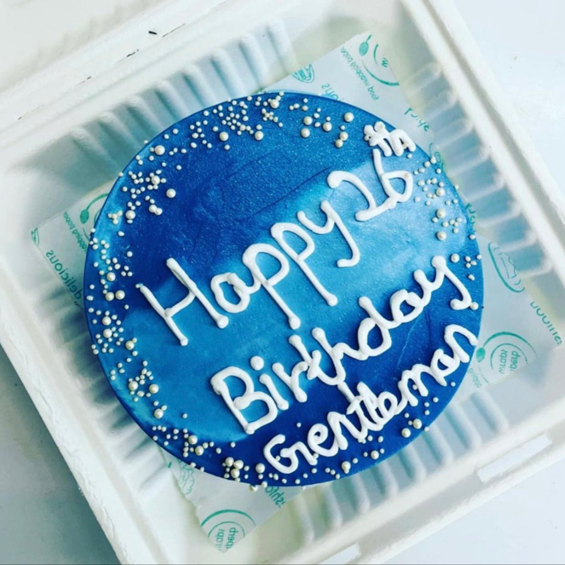 A birthday cake displayed in a clear soft foam container, showcasing its colorful frosting and decorative elements.