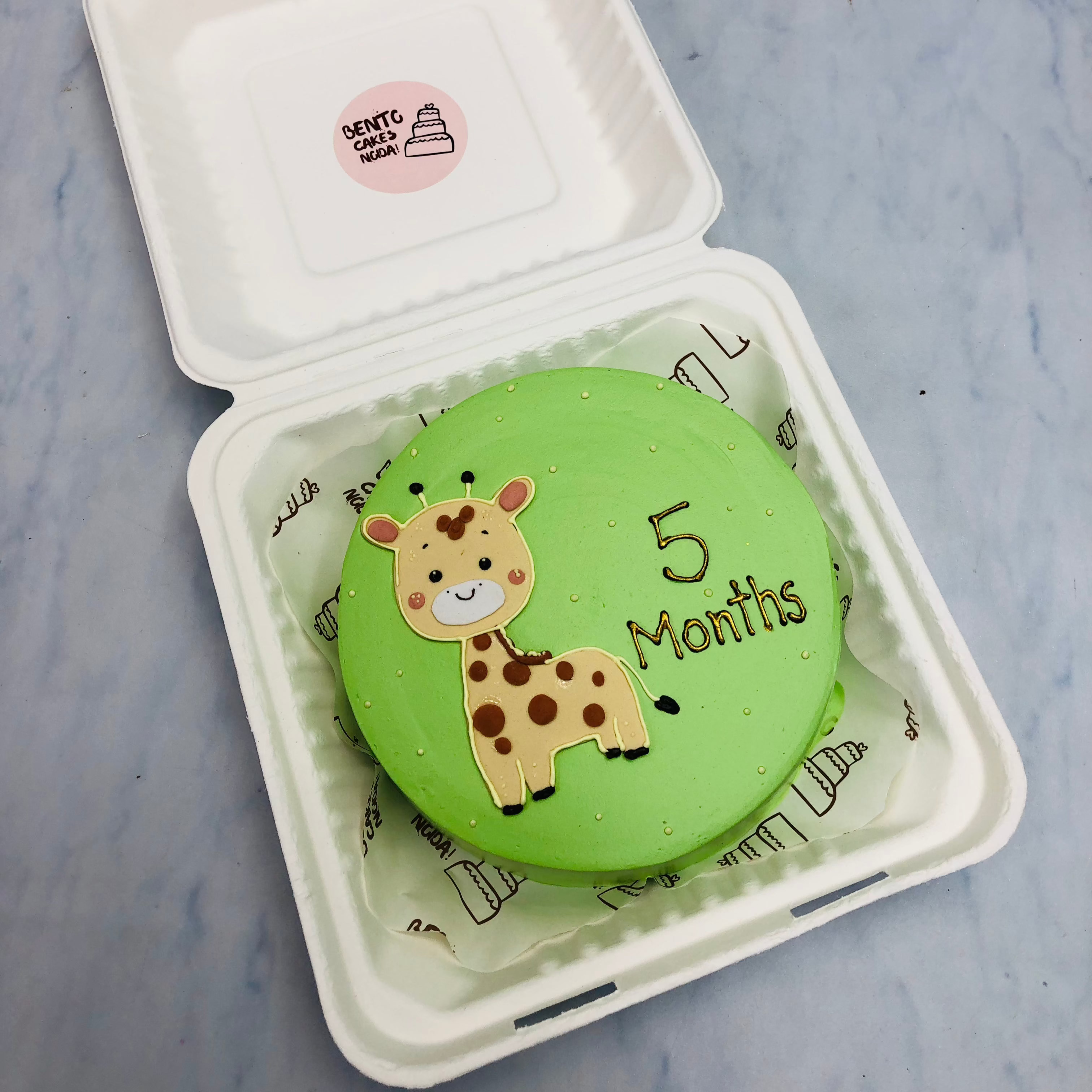 A light green cake in a bento box with a baby giraffe design on it and a 5 months text written with golden color.