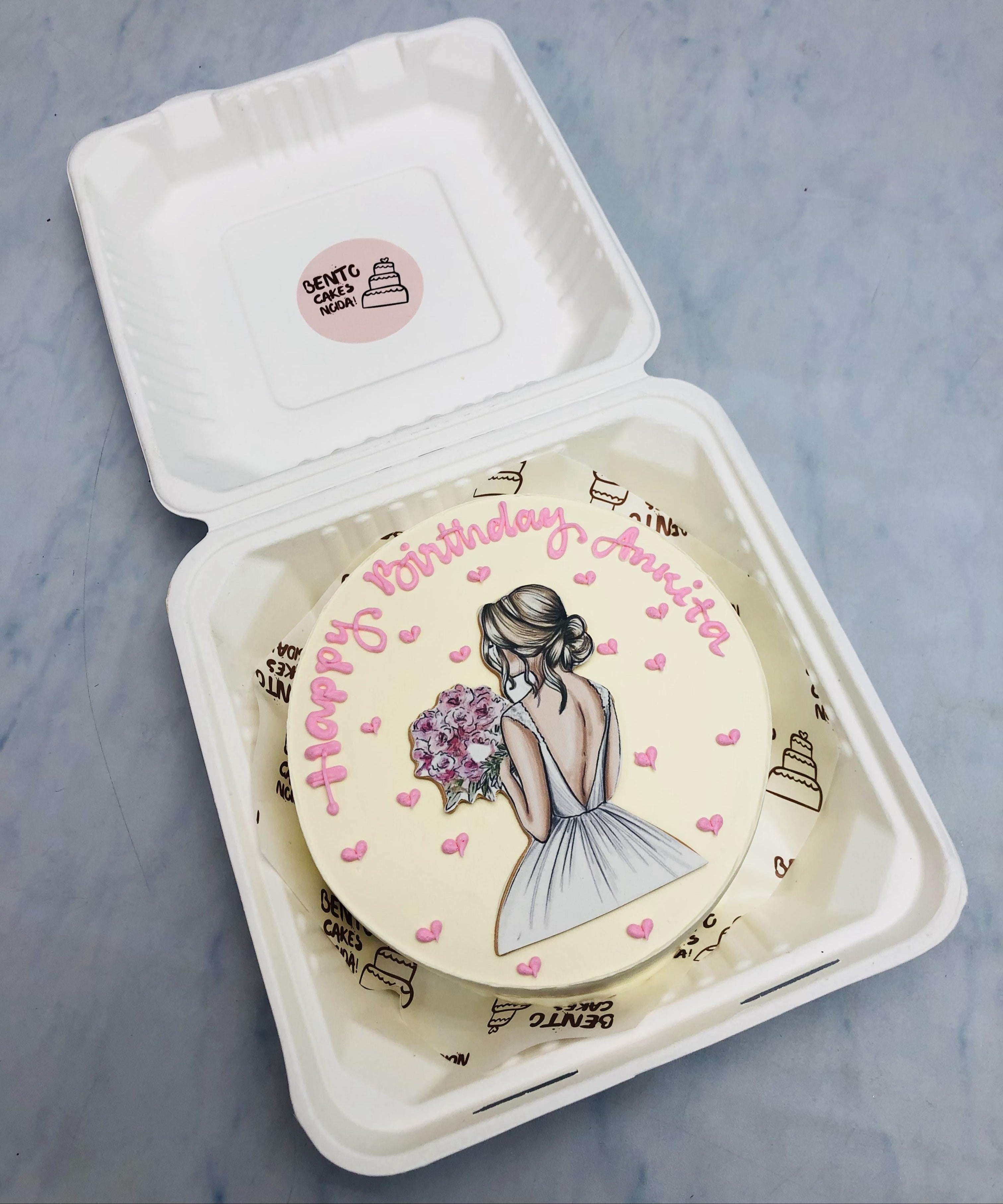 A beautifully crafted birthday cake in a bento box, featuring an artistic illustration of a woman in a white dress holding a bouquet of flowers. Pink heart decorations surround the design, and the message "Happy Birthday Amrita" is elegantly written in pink icing.