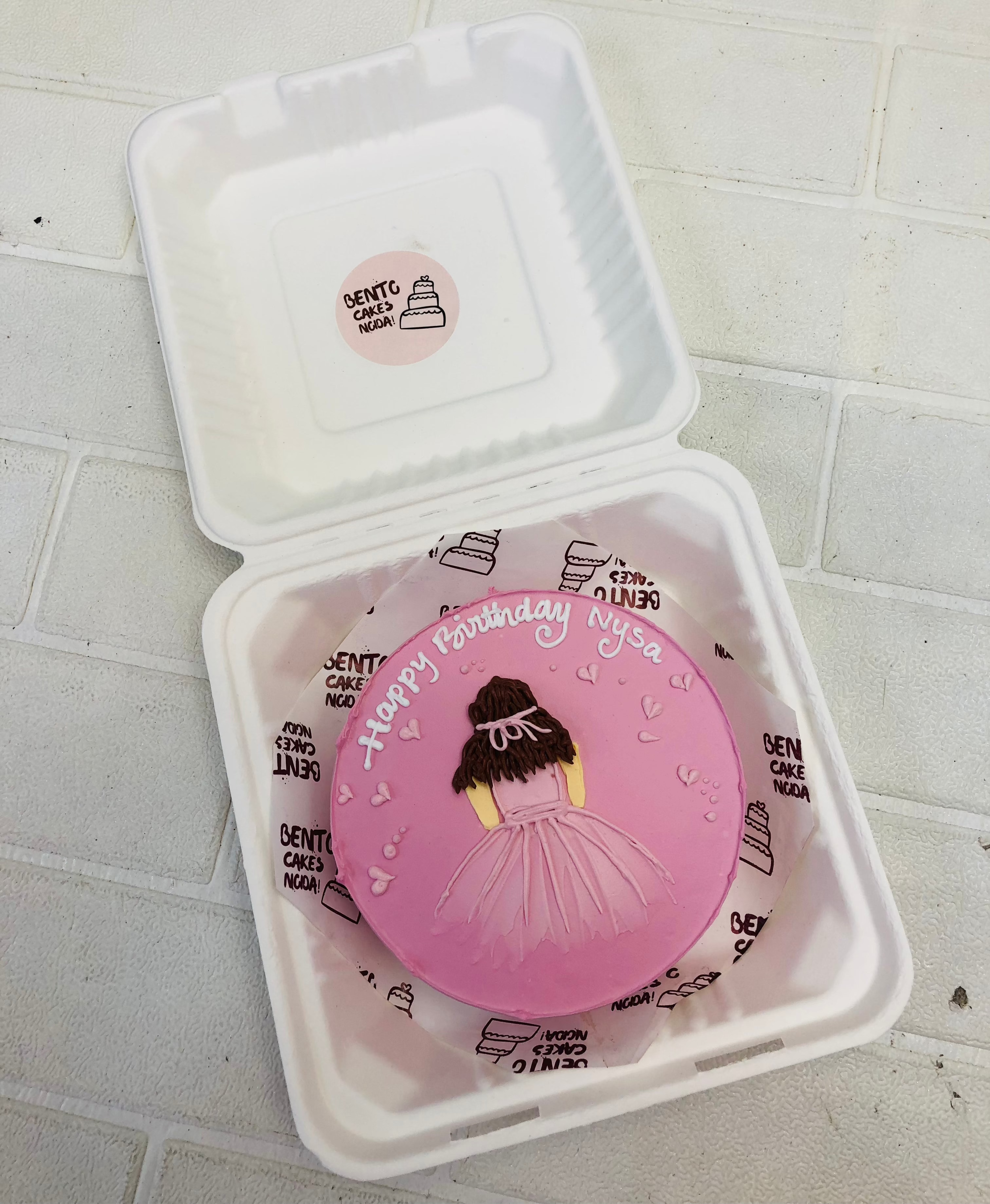 A pink bento cake in a white container with the message "Happy Birthday Nyra" written on top. The cake features a design of a girl in a pink dress with brown hair.