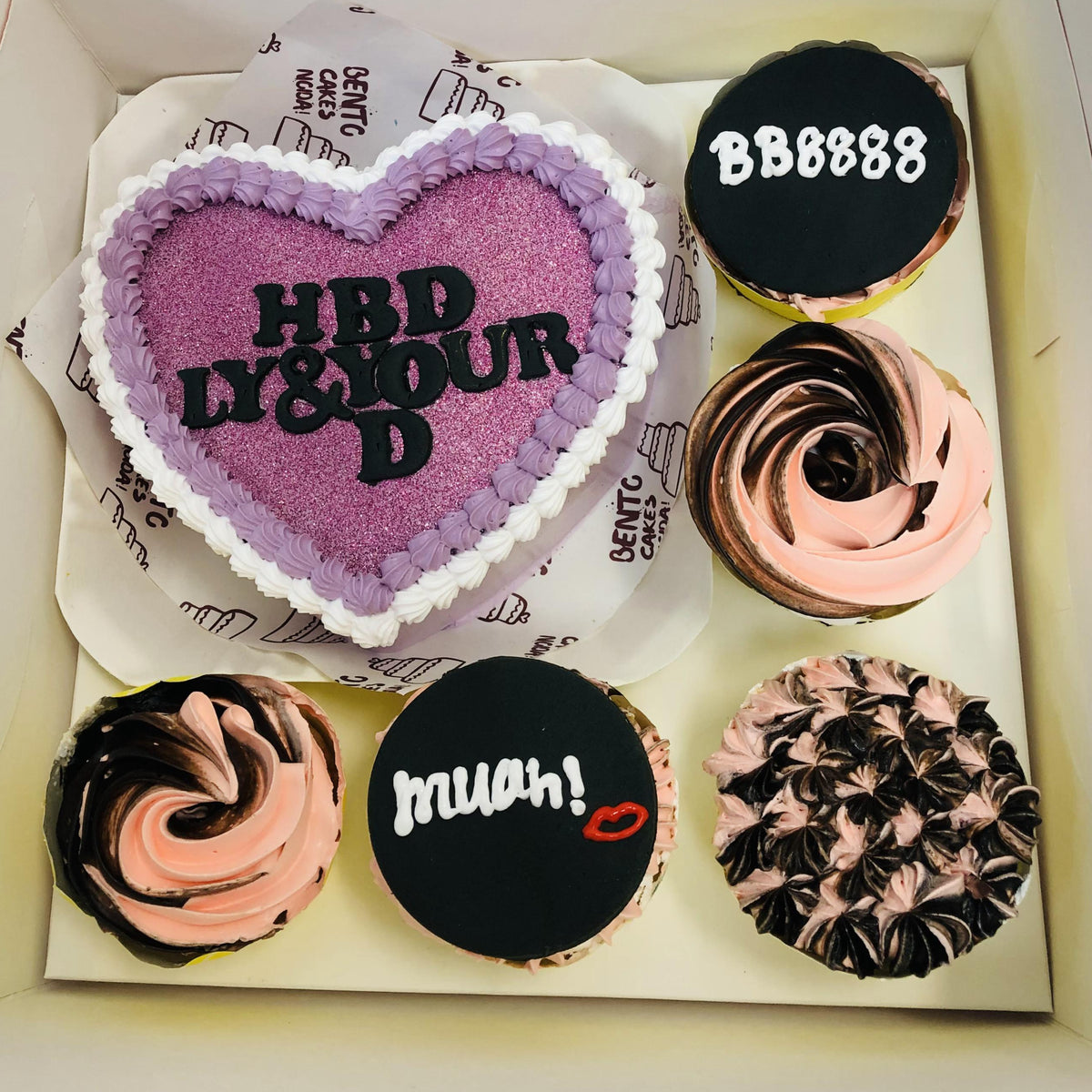 A bento cake set featuring a heart-shaped cake with purple glitter and the text 'HBD LY & YOUR D,' surrounded by five cupcakes with decorative designs including texts 'BB8888' and 'muah!' with a lips motif, and swirl patterns in pink and brown frosting.