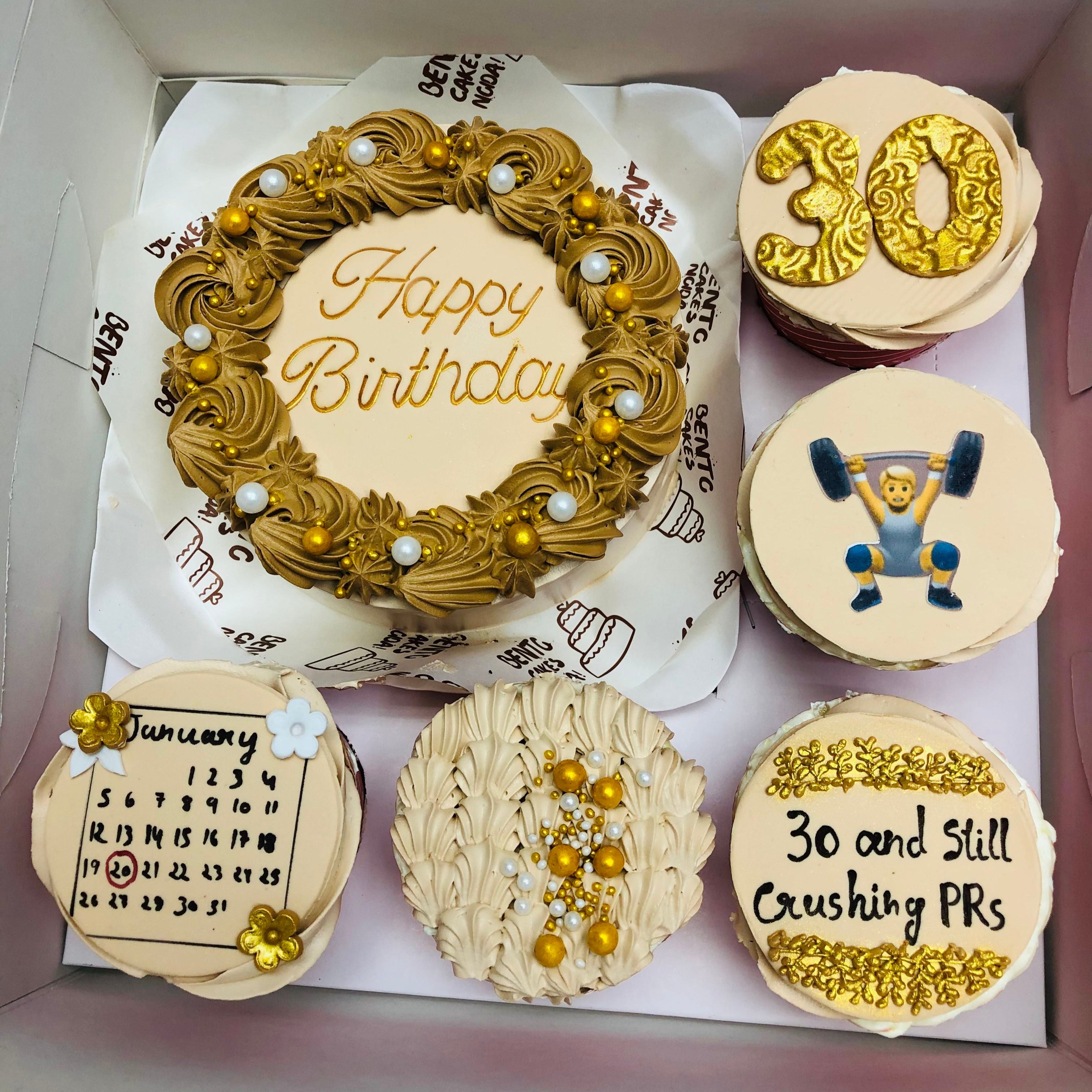 A beautifully decorated birthday cake and cupcakes set. The main cake has "Happy Birthday" written in elegant script, surrounded by intricate frosting and golden decorations. The cupcakes feature various designs, including the number "30" in gold, a weightlifter illustration, a January calendar with a date marked, and the phrase "30 and Still Crushing PRs."