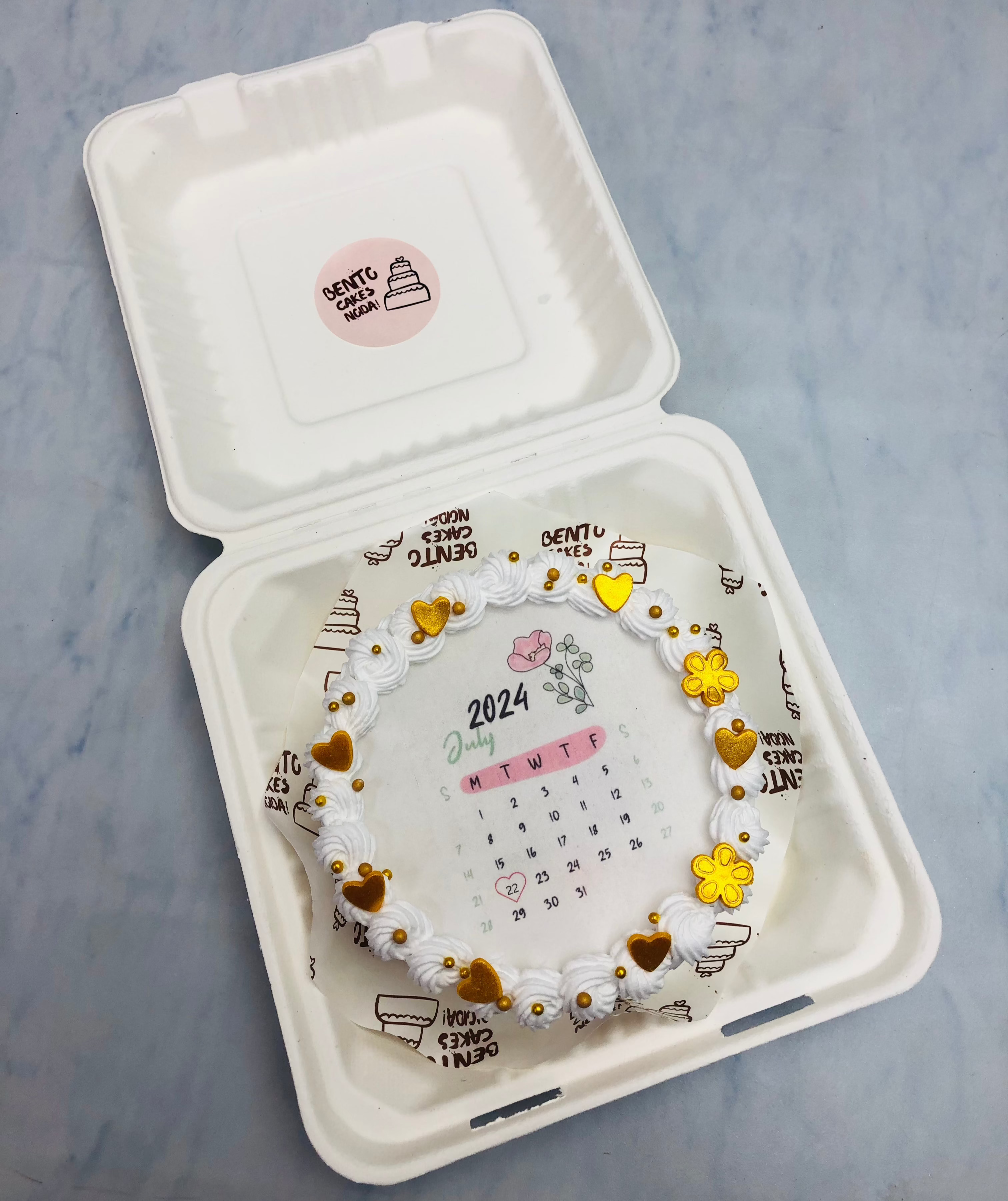 A white truffle fire bento cake decorated with golden hearts and sprinkles on its boundary.