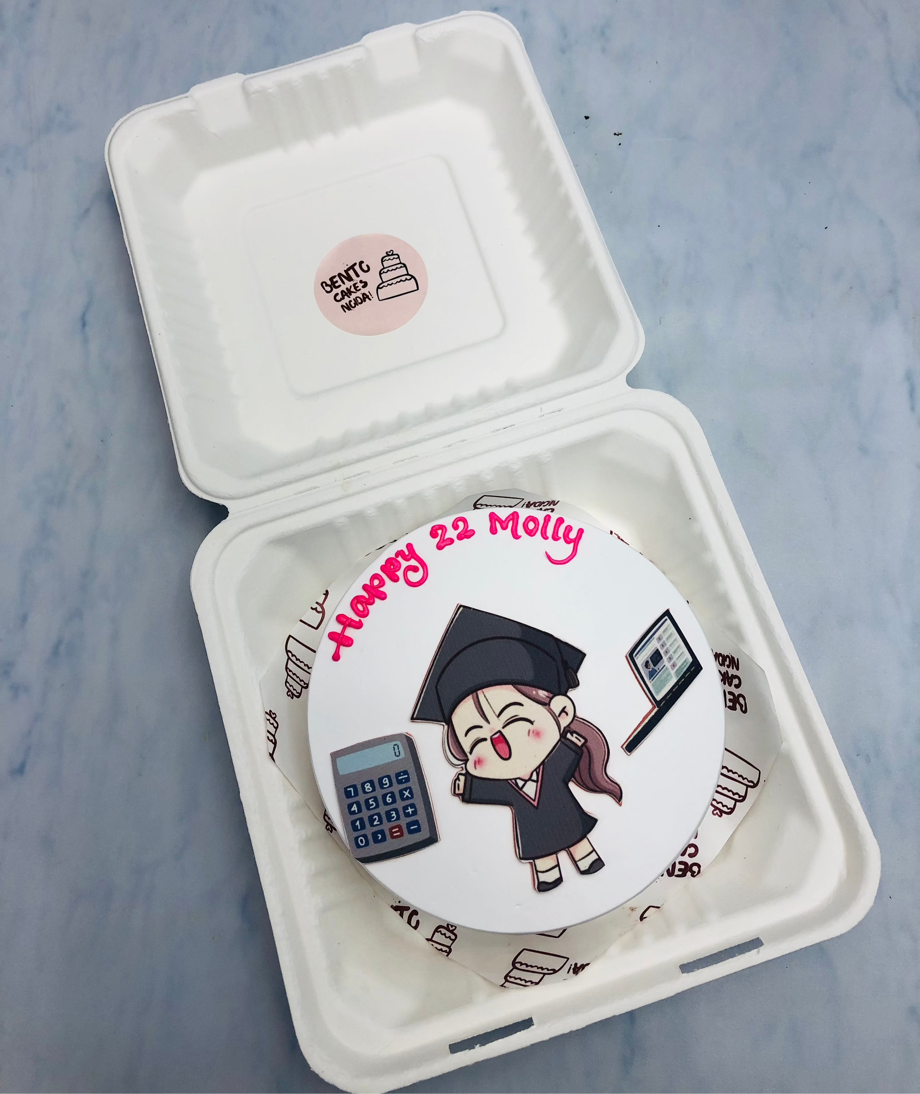 A custom cake in a white box with a cartoon graduate girl, a calculator, and a laptop, with the text 'Happy 22 Molly' written in pink icing.
