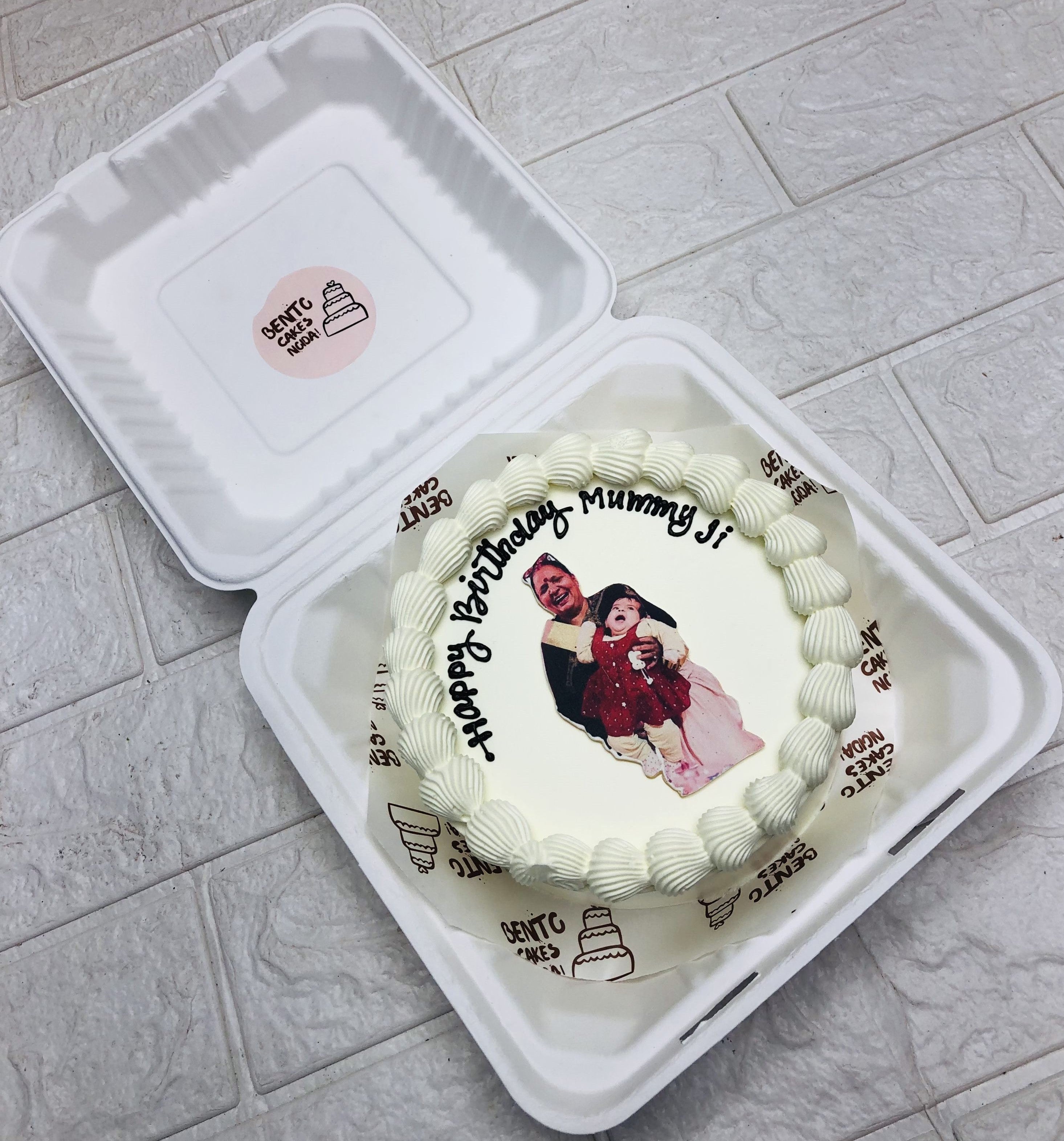 A designer white bento cake having a Grandmother with her grandaughter photo and a "Happy Birthday Mummuy Ji" text written with black icing.