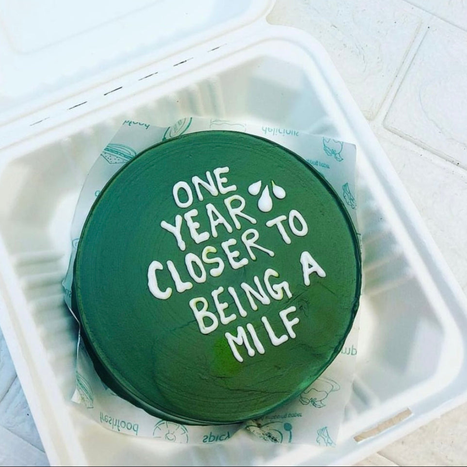 A green cake with white icing text that reads "ONE YEAR CLOSER TO BEING A MILF" inside a white takeout container.