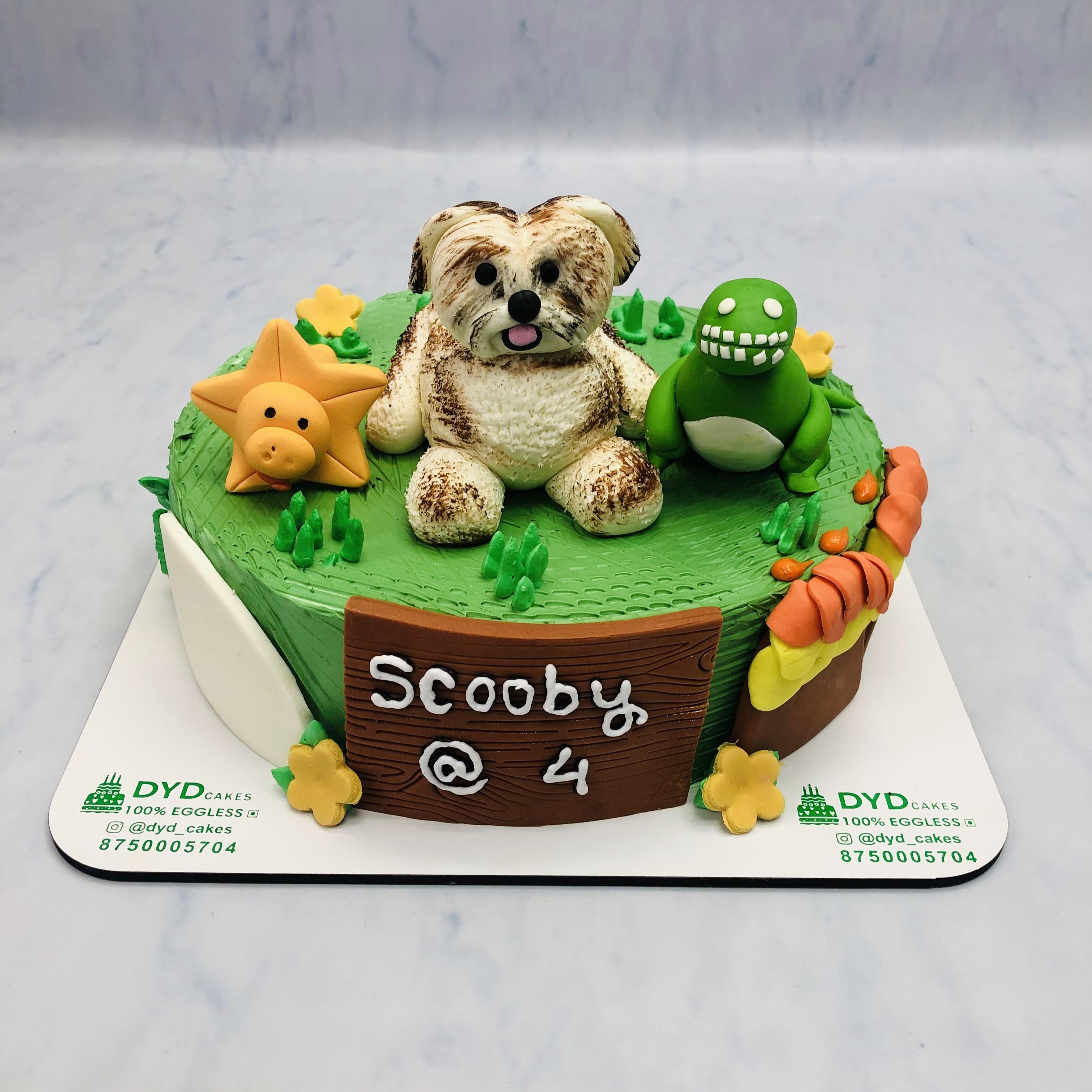 A green birthday cake with a dinosaur and star decoration, featuring the text 'Scooby @ 4' on a wooden sign.